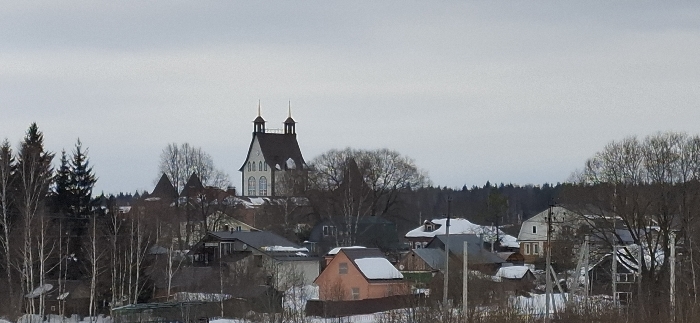 This image depicts a panoramic view of a settlement identified as Minino, Russia, under an overcast sky. The foreground and midground feature a collection of residential-style buildings interspersed with bare deciduous trees and some evergreen conifers. Patches of snow cover the ground and many rooftops, indicating a winter or early spring season.
A prominent, light-colored structure with a dark, multi-gabled roof and two distinct spires rises on an elevated position towards the center-left, featuring multiple arched windows. Surrounding this central building are numerous detached houses of varying sizes and architectural styles, predominantly one to two stories, with a mix of dark and lighter-colored roofs, several of which bear a layer of snow.
Vegetation consists primarily of leafless deciduous trees with visible branches, creating a skeletal appearance, alongside darker coniferous trees. A dense line of forest forms the distant horizon across the upper right and left portions of the image. No individuals or specific actions are discernible within the frame. The overall environment suggests a quiet, rural or semi-rural setting.