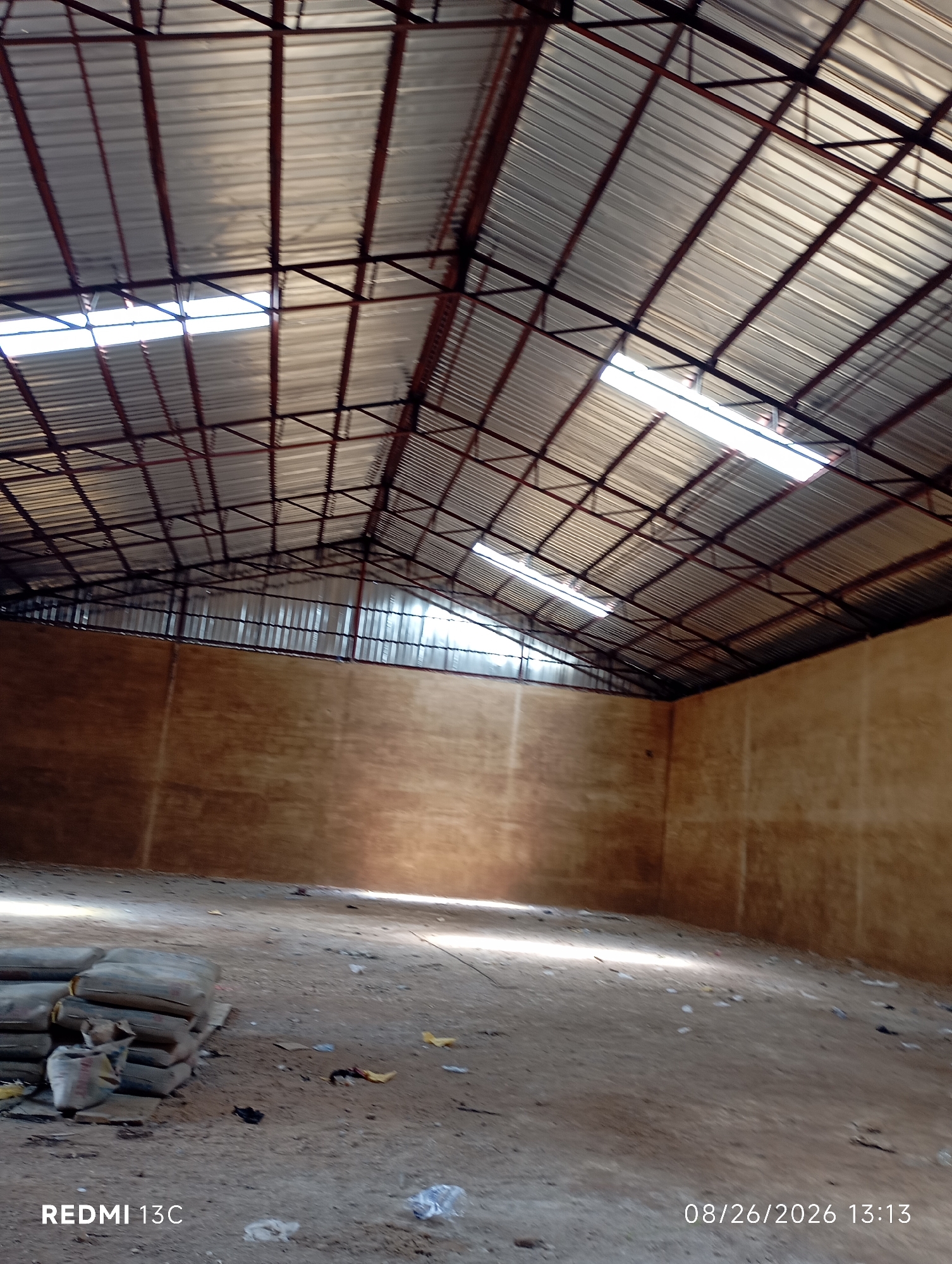 The image depicts the interior of a large, empty warehouse located in Shendam, Nigeria. The warehouse has a corrugated metal roof supported by a dark metal frame, with several skylights allowing natural light to enter. The walls appear to be made of a light brown, possibly concrete or plaster material. The floor is dirt or concrete, littered with some debris, including plastic bags and yellow paper. In the foreground, on the left side of the image, a stack of grey sacks, possibly containing cement, is visible. The overall impression is of an unfinished or disused industrial space. The bottom of the image contains a watermark indicating "REDMI 13C" and the date "08/26/2026 13:13". There are no people or animals present in the scene.