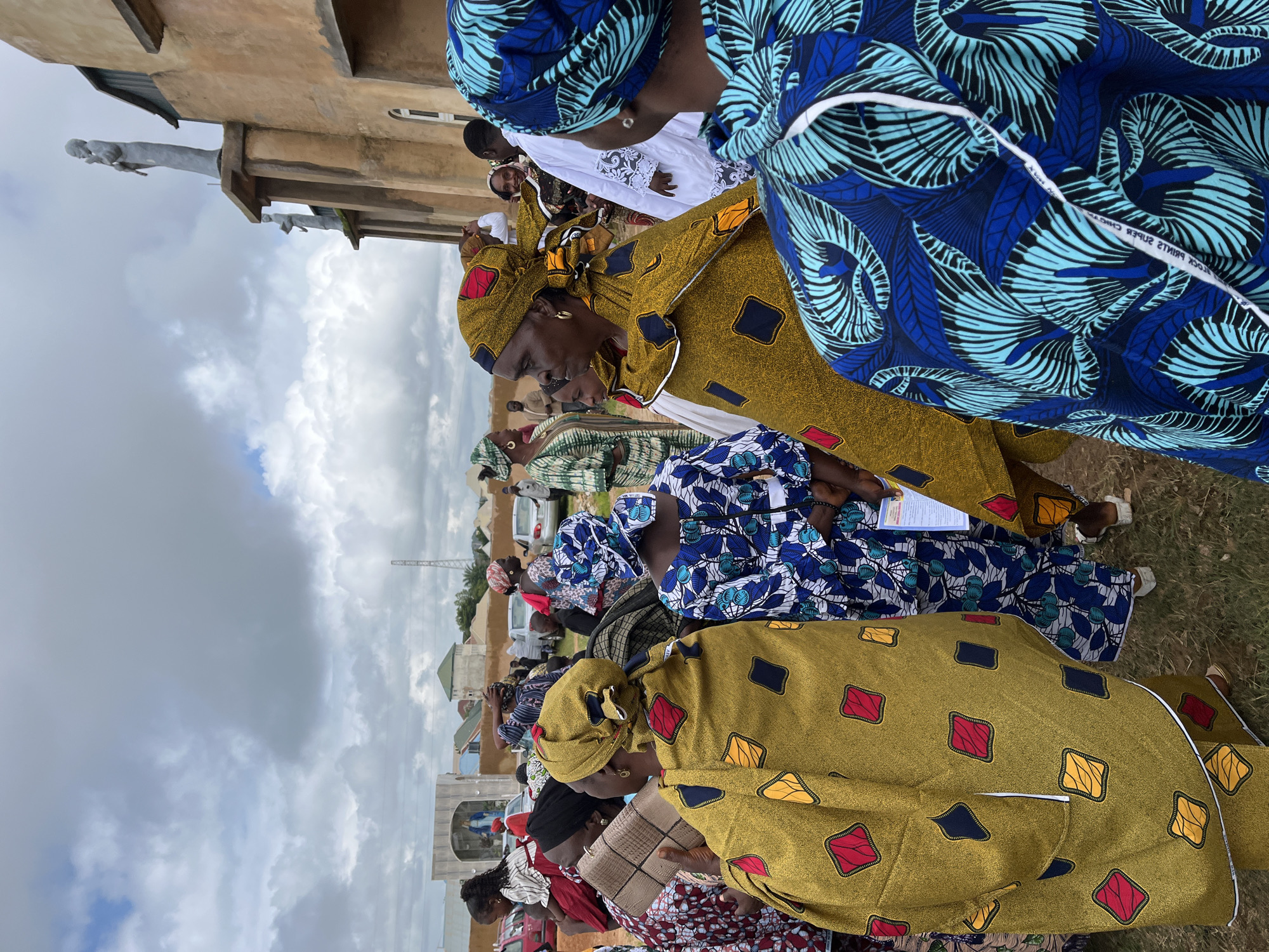 An outdoor medium shot captures a group of people, predominantly women, gathered near a building adorned with statues. The scene appears to be set in Jos, Nigeria, based on the context of the query.
The foreground is dominated by several women wearing brightly colored, patterned traditional African attire. One woman in a mustard yellow garment with square red, yellow, and black patterns is facing away from the camera, her head turned slightly to her left. Another woman to her right wears a blue and white patterned outfit, also with her back mostly to the viewer. To the left, a woman in a maroon and white striped dress is partially visible.
In the background, more people are milling about. A building with a tan facade and visible architectural details, including statues on its upper level, is prominent. The sky above is cloudy, with patches of blue peeking through. The ground appears to be covered in dry grass. Several vehicles are parked in the middle ground, suggesting a gathering or event. The overall atmosphere seems social and communal.
