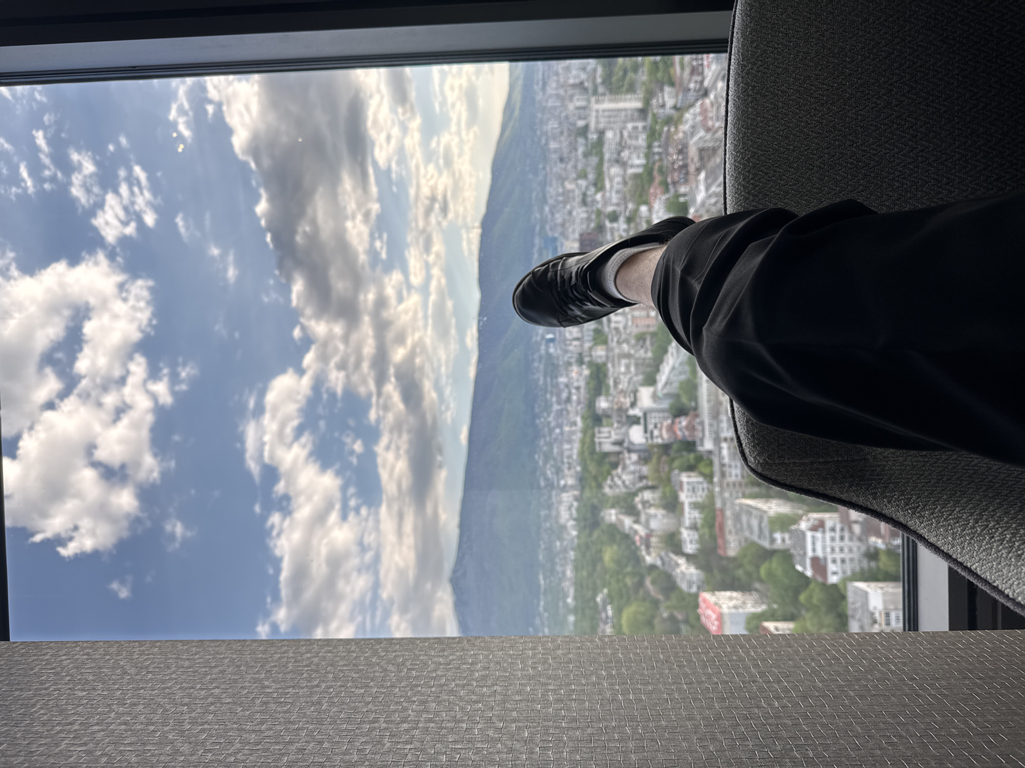 A view from a high-rise building in Sofia, Bulgaria, looking out a window. Visible through the window are: a cloudy blue sky, the outline of Vitosha Mountain, and a cityscape with residential and commercial buildings interspersed with trees. Part of a chair and a person's leg, clad in black pants and a black shoe, are visible on the right. The interior surface in the foreground is a textured gray material.