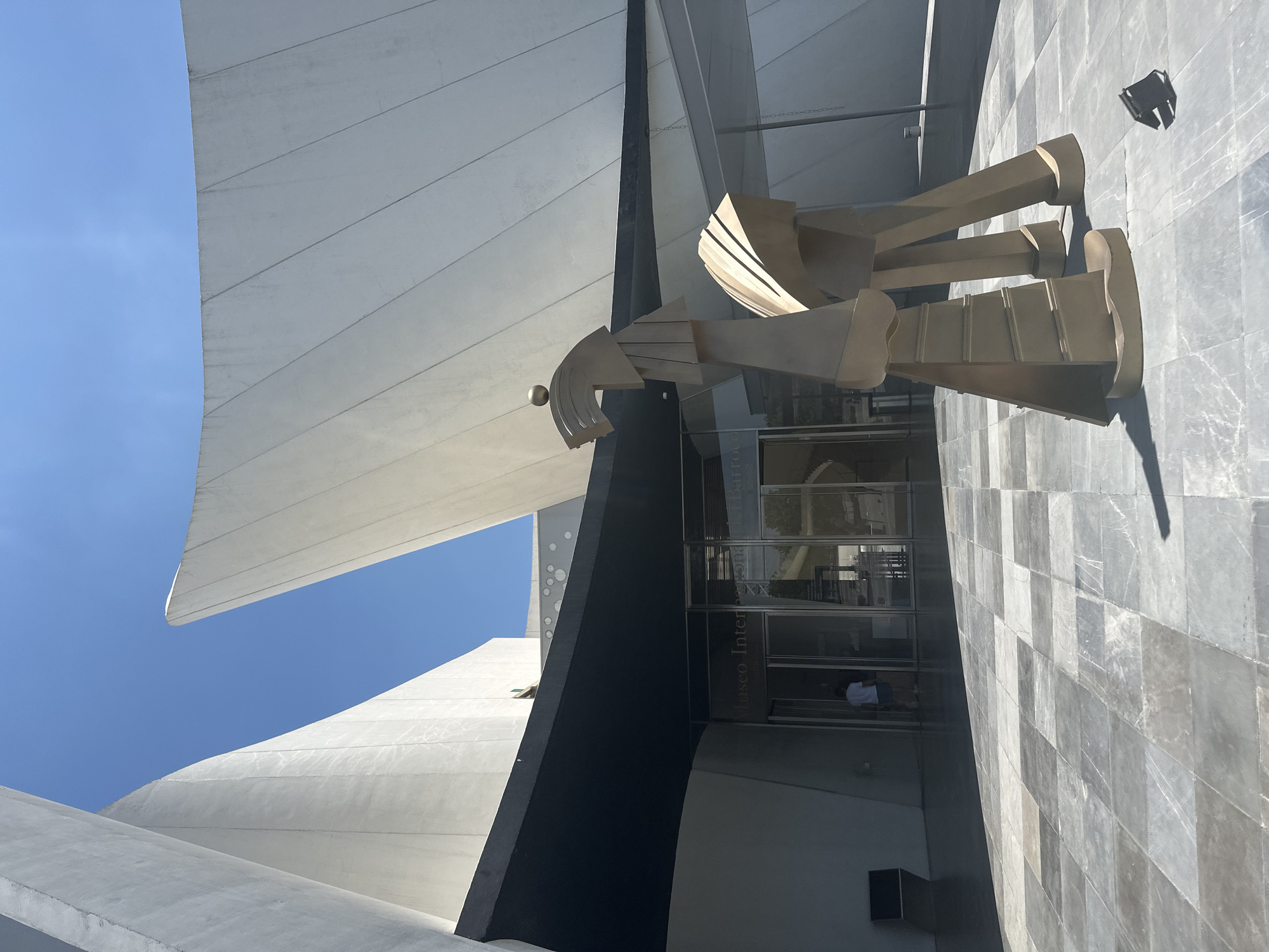 This media file captures an exterior view of a modern architectural complex under a clear, bright blue sky. The scene is dominated by striking, sweeping white structures that resemble large sails or wings, contrasting with angular dark grey or black building elements.
In the foreground, a large, abstract sculpture made of geometric, light-colored metal components is prominently displayed. The sculpture features distinct shapes that evoke a figure, with what appear to be legs and feet on a tiled ground, and an intricate, angular upper body extending towards the building's facade.
The building itself features a glass entrance, through which the words "Museo Inter" are visible, suggesting it is an "International Museum." Inside the entrance, a person in a white shirt can be faintly seen, possibly a staff member. The ground and a wall to the right are clad in a checkerboard pattern of grey and lighter grey tiles.
The bright sunlight indicates it is daytime, casting sharp shadows from the sculpture and architectural features onto the ground and walls, emphasizing the geometric forms and textures of the modern design. The overall impression is one of contemporary architecture, art, and an open, light-filled environment, consistent with a cultural institution.