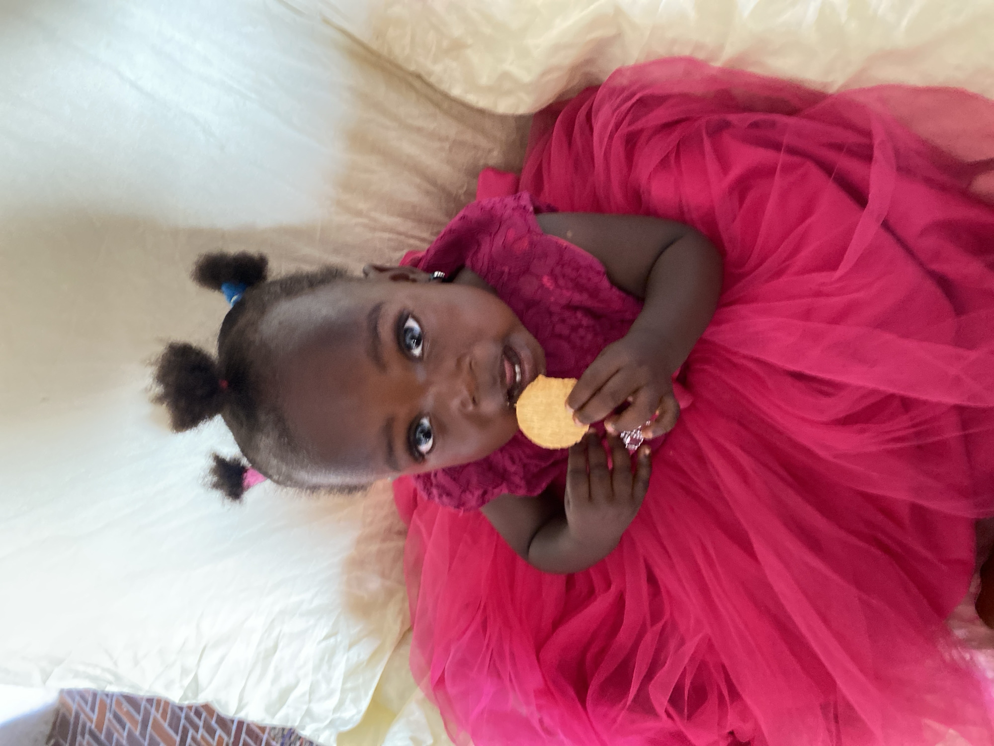 A young child, likely a toddler, is captured in a high-angle shot, lying down on a light-colored sheet or blanket. The child has dark skin and dark hair styled into two small pigtails, one with a blue hair tie and the other with a pink hair tie. They are wearing a bright fuchsia-colored dress, featuring a layered, voluminous skirt made of a sheer, tulle-like fabric and a textured bodice.
The child looks directly at the camera with wide eyes and an open mouth, appearing to be in the midst of eating. They hold a round, light-colored biscuit or cookie in their right hand, close to their mouth. A shiny ring is visible on the ring finger of their left hand.
The setting appears to be indoors in Vom, Nigeria, with a small portion of a tiled floor, featuring brown and tan brick patterns, visible in the lower left corner of the frame. The child's expression conveys curiosity and engagement, possibly capturing a moment of playful snacking. No text is visible in the image.