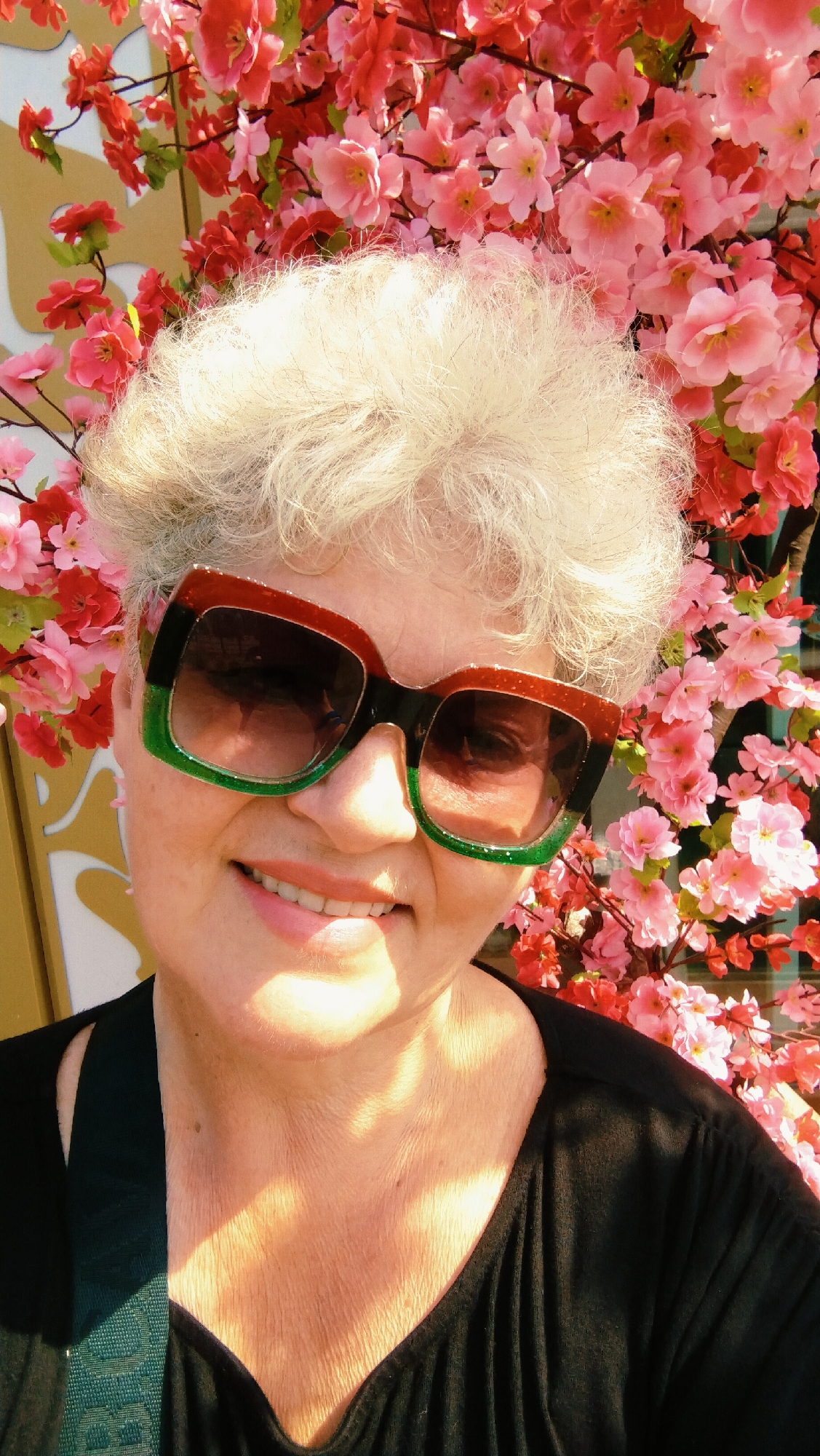 A close-up, over-the-shoulder selfie shows an older woman with short, white, voluminous hair and fair skin, wearing large, angular sunglasses with green and reddish-brown frames. She is smiling, revealing her teeth and rosy lips, and her head is tilted slightly to the left. The woman is wearing a black top, with a dark green strap visible over her left shoulder. Behind her, a backdrop of vibrant pink and red artificial cherry blossoms is prominent. The lighting suggests an outdoor setting, with dappled sunlight illuminating her neck and chest. The background also shows a hint of a golden-yellow structure, possibly a building facade or a decorative element, to the left of the woman. The overall impression is of a cheerful portrait set against a colorful, decorative background, likely taken during a sunny day in Horishni Plavni, Ukraine.