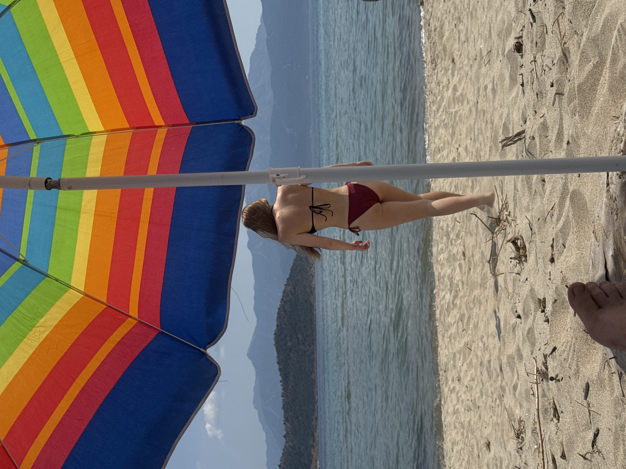 The image captures a beach scene from the perspective of someone seated beneath a large, rainbow-striped beach umbrella. A bare foot is visible in the bottom left foreground, partially buried in the light brown sand which is scattered with natural debris like driftwood and dry vegetation. A light-colored umbrella pole runs vertically in the center, partially obscuring the midground. A female figure, viewed from behind, walks towards the ocean. She has long, light-colored hair and is wearing a black bikini top and maroon-colored bikini bottoms. The ocean water appears calm with gentle waves breaking near the sandy shore. In the background, a tree-covered landmass is visible across the water, backed by a range of hazy, distant blue mountains under a mostly clear sky with a few sparse white clouds. The location is Unknown, Unknown.