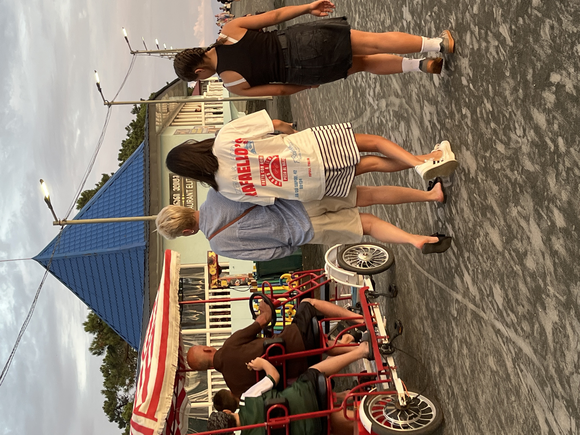 In Ureki, Georgia, at sunset, a group of people are seen from behind on a paved walkway. In the foreground, a red and white striped canopy covers a multi-person pedal cart, occupied by at least two individuals. A man with a bald head is seated at the rear, operating the steering wheel. Beside him, a person wearing a green vest sits on the cart. To the right of the cart, three individuals walk away from the camera. The first person on the left wears a blue and white striped long-sleeved shirt and tan shorts. Next to them is a person in a white t-shirt with "RAFAELLO'S" printed in red and a striped skirt. The third person is a young female in a black tank top and a denim skirt. In the background, a building with a blue, tiled, pyramid-shaped roof is visible, along with trees and lampposts lining the walkway. The sky is overcast with hints of warm light from the setting sun.