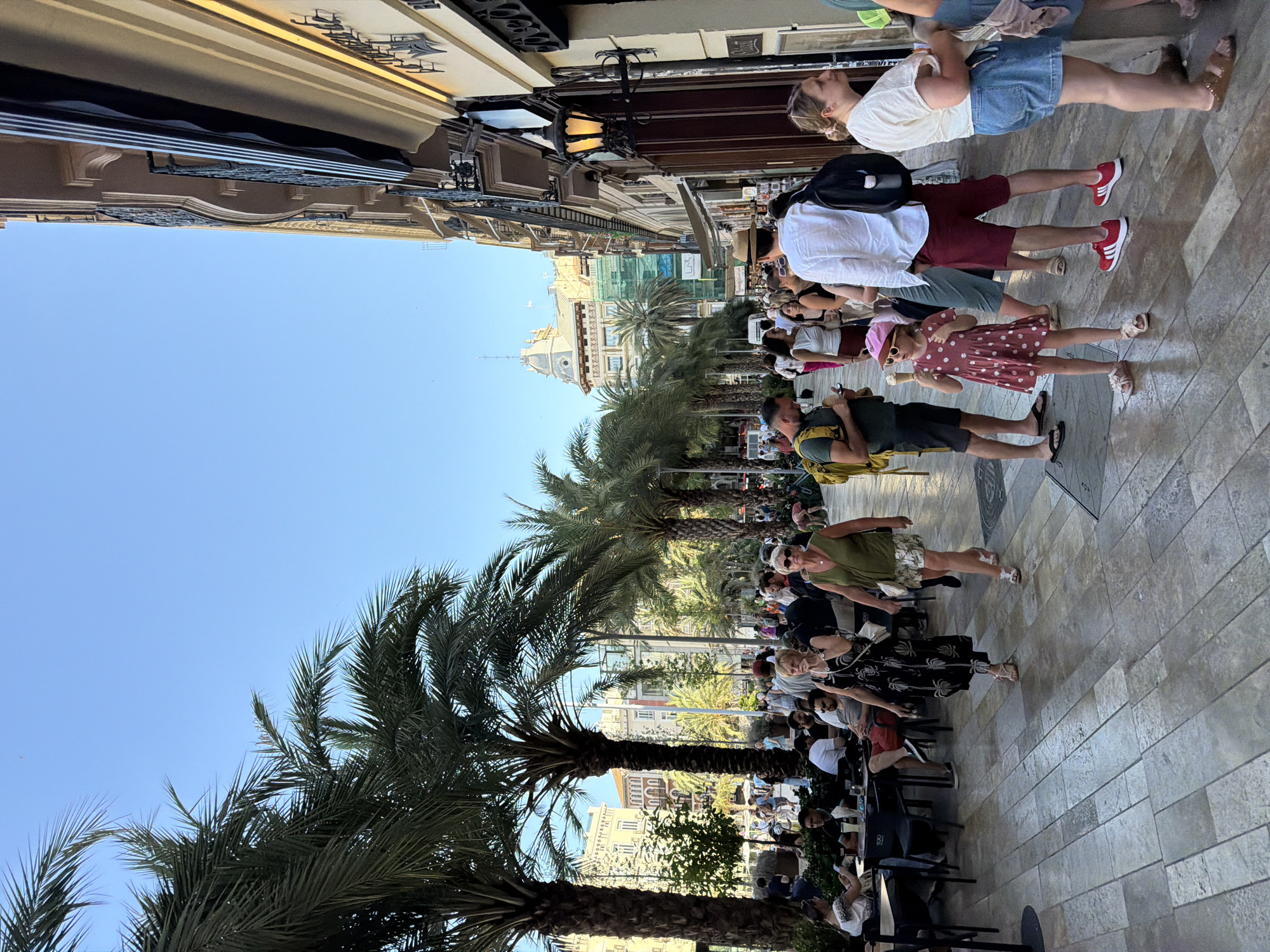 In an outdoor urban setting, a wide, paved walkway lined with palm trees and buildings is bustling with pedestrians. The sky is clear blue. On the left, several people are seated at tables, seemingly at an outdoor cafe. A woman in a black floral dress and sandals walks toward the right, followed by a woman in a green sleeveless top and a patterned skirt. Further to the right, a young girl in a pink polka dot dress and a pink hat holds an ice cream cone, and a man in a white shirt and straw hat walks past her. Men and women in casual summer attire populate the scene. The buildings on the right feature architectural details like balconies and awnings, with a lit lantern on an exterior wall. In the background, more buildings are visible, some under construction with scaffolding.