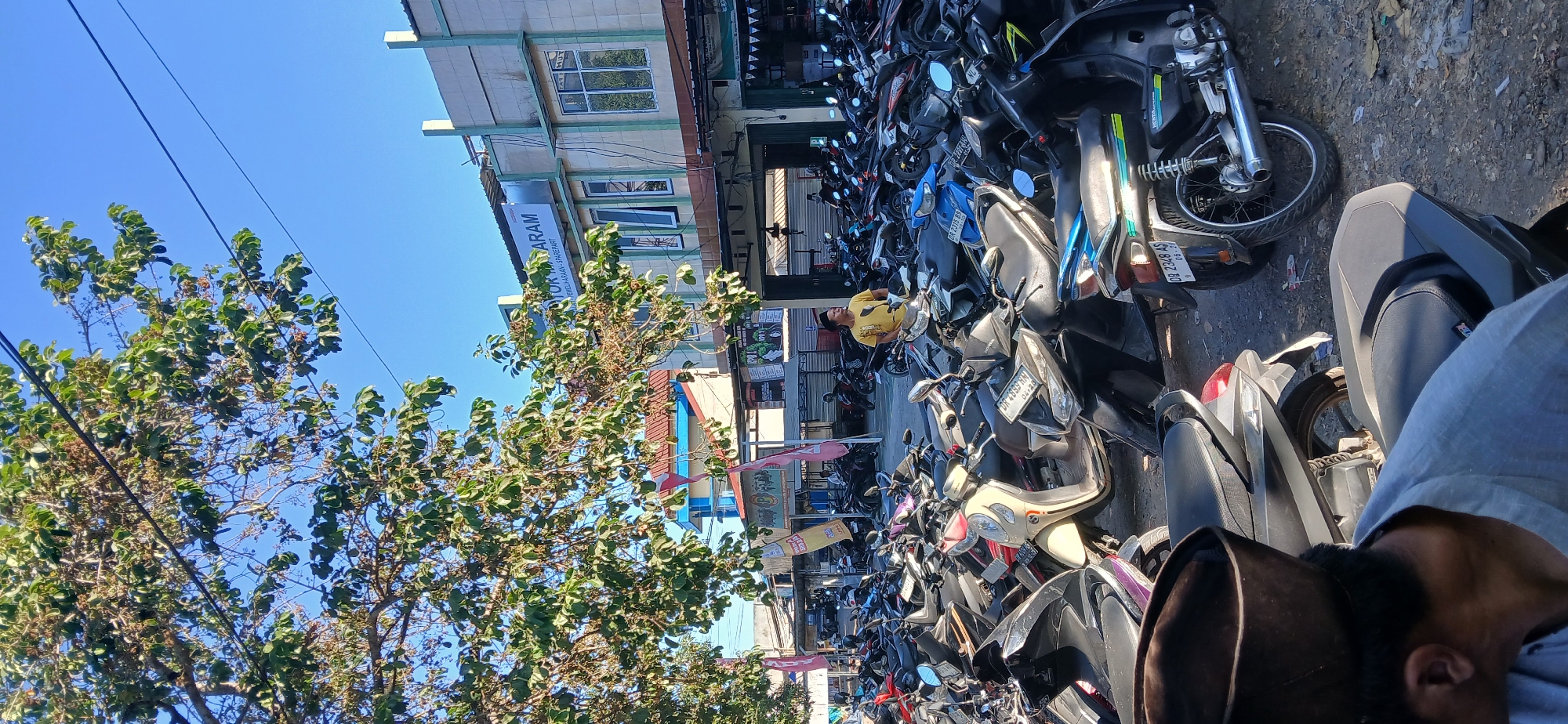 A photograph captured from a low angle shows a densely packed parking lot filled with numerous motorcycles under a bright, clear blue sky. The foreground features the back of a person wearing a light blue shirt and a dark brown cap, partially obscuring some motorcycles. In the mid-ground, a man in a yellow t-shirt stands near a parked motorcycle, looking towards the right. Behind him, a two-story building with an awning extends across the front. The building's facade is light beige with green accents around the windows and roofline. A sign above the entrance reads "DIKTOR MASARAM" followed by smaller text in Indonesian. The environment appears to be an urban setting in Kediri, Indonesia, likely a commercial area or a public parking facility. The lighting suggests it is daytime, with strong sunlight casting shadows. The overall scene depicts a busy, everyday street view in the city.