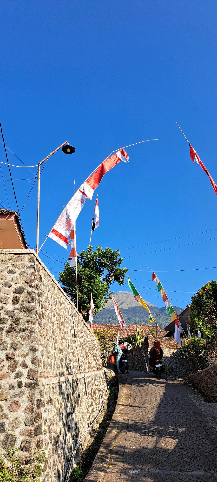The image shows a steep, paved alleyway in Sukoreno, Indonesia, lined with stone walls. Several Indonesian flags, predominantly red and white, are displayed on tall poles, some of which are extended horizontally by wind. The flags are of various sizes and designs, with some featuring intricate patterns. In the background, a lush green tree stands tall, and beyond it, a mountain peak is visible under a clear blue sky. Two individuals on motorcycles are proceeding up the alleyway. The left motorcycle has a passenger wearing a turquoise shirt and carrying a red bag. The right motorcycle has a passenger wearing a brown headscarf and a dark shirt. The overall scene depicts a vibrant, sunlit day in a rural Indonesian setting.
