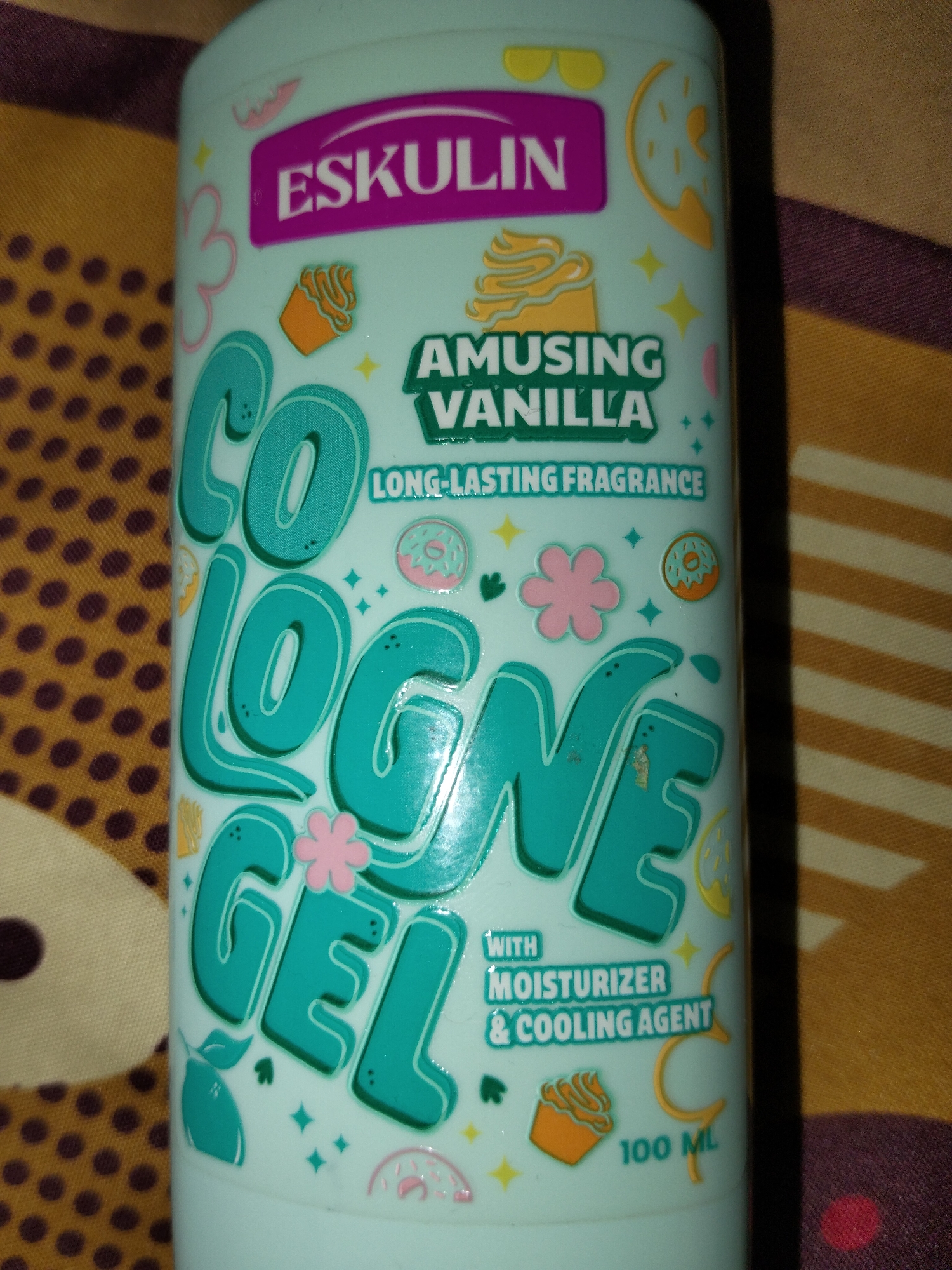A light blue bottle of Eskulin Cologne Gel with vanilla fragrance is shown, positioned vertically. The bottle features various decorative elements, including stylized cupcakes, donuts, and flowers, printed in pink, orange, and yellow. Prominently displayed are the words "ESKULIN" in large pink letters, followed by "AMUSING VANILLA" in green text, and then "COLOGNE GEL" in large turquoise letters. Below this, smaller text reads "LONG-LASTING FRAGRANCE" and "WITH MOISTURIZER & COOLING AGENT". The bottom of the bottle indicates "100 ML". The background consists of a patterned fabric with alternating stripes of yellow and orange. The image is taken indoors, and there are no people or animals visible.