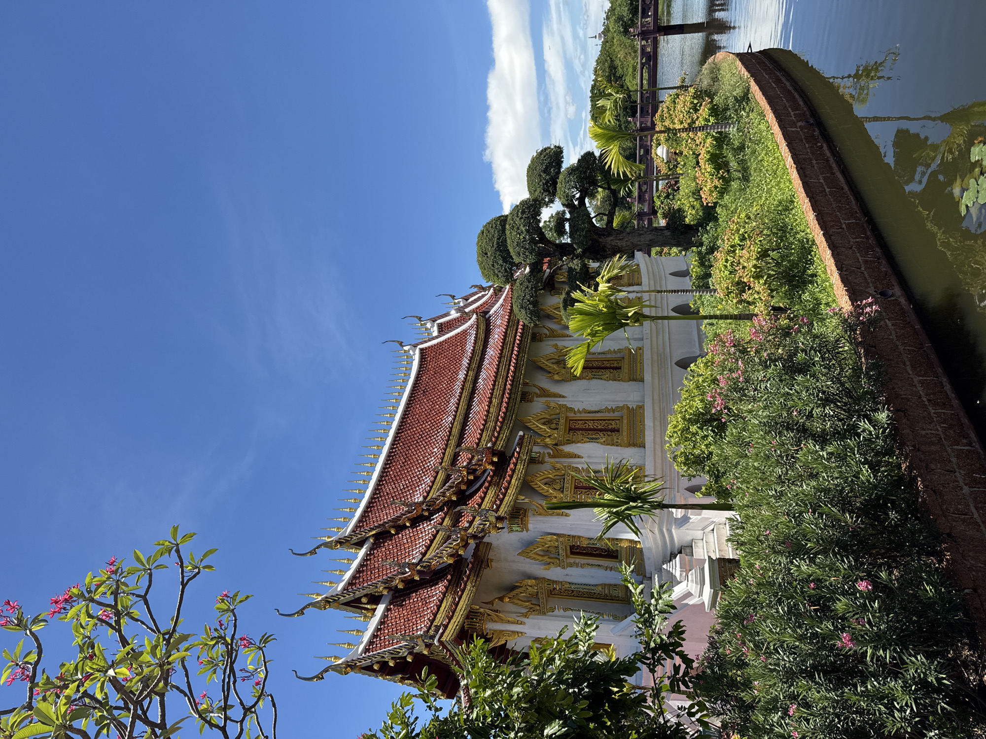 The image depicts a temple structure in Bang Pu Subdistrict Municipality, Thailand, characterized by a traditional multi-tiered red and gold roof. The temple is surrounded by lush greenery including decorative trees, shrubs with pink flowers, and palm trees. A body of water with a brick-lined edge is adjacent to the vegetation, reflecting the sky. The sky is blue with scattered clouds. A bridge is visible in the background over the water.