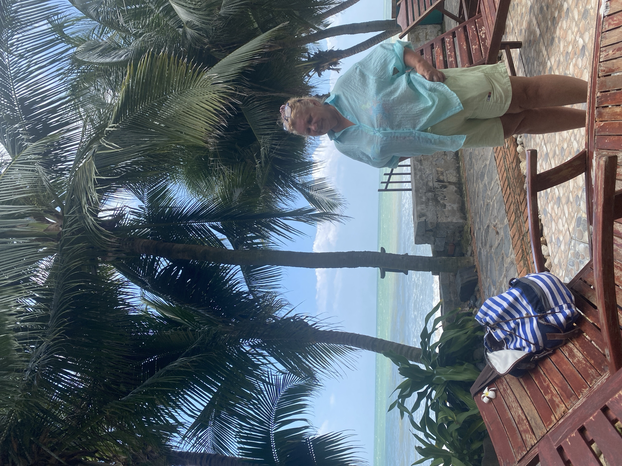 A woman stands on a tiled patio in Mũi Né, Vietnam, with palm trees and the ocean in the background. The woman is wearing a light blue, translucent button-up shirt and light green shorts. She is looking down and to her left. In front of her, to the left, is a wooden lounge chair with a blue and white striped backpack and a white flower on it. Behind her, to the right, is another wooden lounge chair. The ocean is visible beyond a low stone wall, with gentle waves breaking on the shore. The sky is blue with scattered clouds.