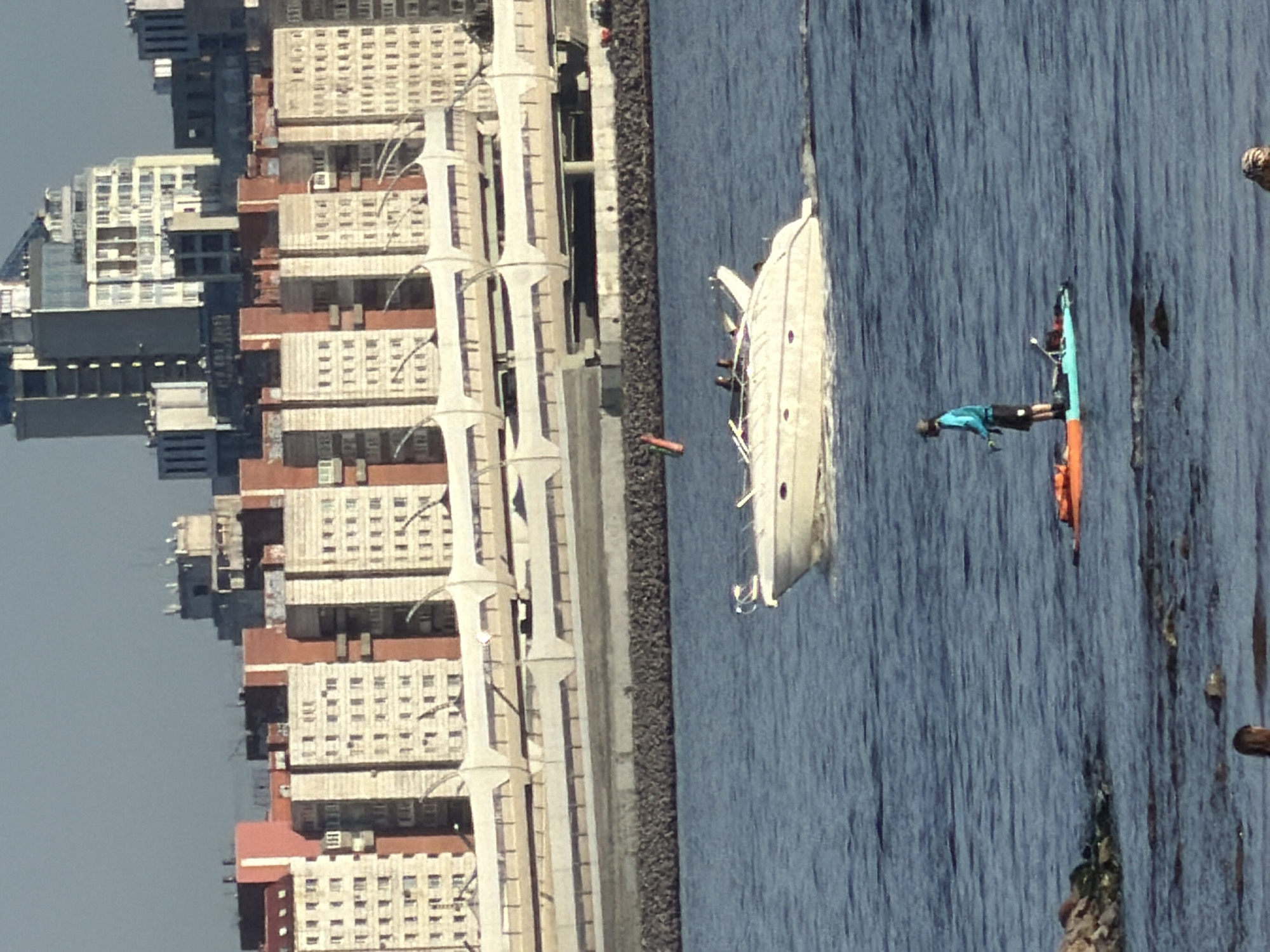 The image depicts an urban waterfront scene in Saint Petersburg, Russia, under bright daylight. A wide expanse of calm, deep blue water occupies the foreground and midground. On the water, a white recreational yacht or boat is prominently featured, with a few indistinct figures visible on deck. To the right of the yacht, a person in a blue top and black shorts is actively engaged in stand-up paddleboarding, balancing on a blue and green paddleboard. A second, orange paddleboard is partially visible nearby, along with a small red buoy or float in the water closer to the shore.
A long, grey concrete or stone embankment separates the water from the city. Behind this embankment, a densely packed cityscape rises under a clear, light grey sky. Numerous multi-story buildings, likely residential or commercial, line the waterfront. These structures feature a mix of white facades with many windows and sections of reddish-brown. Distinctive, tall, light-colored, column-like architectural elements are visible along the embankment, possibly belonging to a large complex or a bridge. The scene suggests a pleasant day for outdoor and water-based recreational activities within a bustling urban environment. No discernible text is visible in the image.