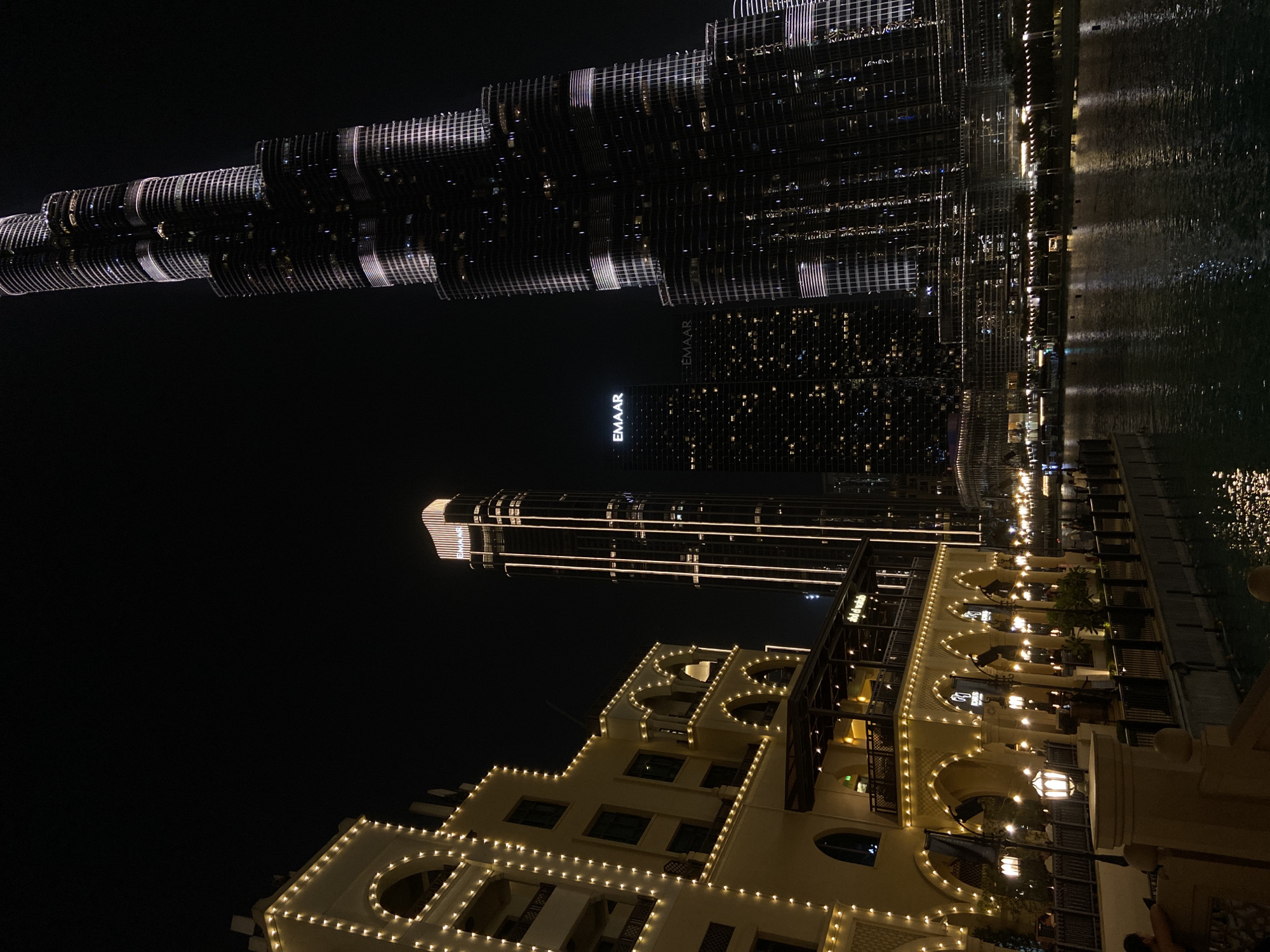 The image depicts a night scene in Dubai, United Arab Emirates, featuring the Burj Khalifa and adjacent structures. The Burj Khalifa, a prominent skyscraper, is visible at the top. Its illuminated facade is reflected in the water. Below the Burj Khalifa is another skyscraper. The building in the foreground is adorned with lights and contains archways and balconies. The image is taken at night, and the lighting emphasizes the architecture of the buildings. There are no people explicitly visible in the image, and the focus is on the urban landscape.