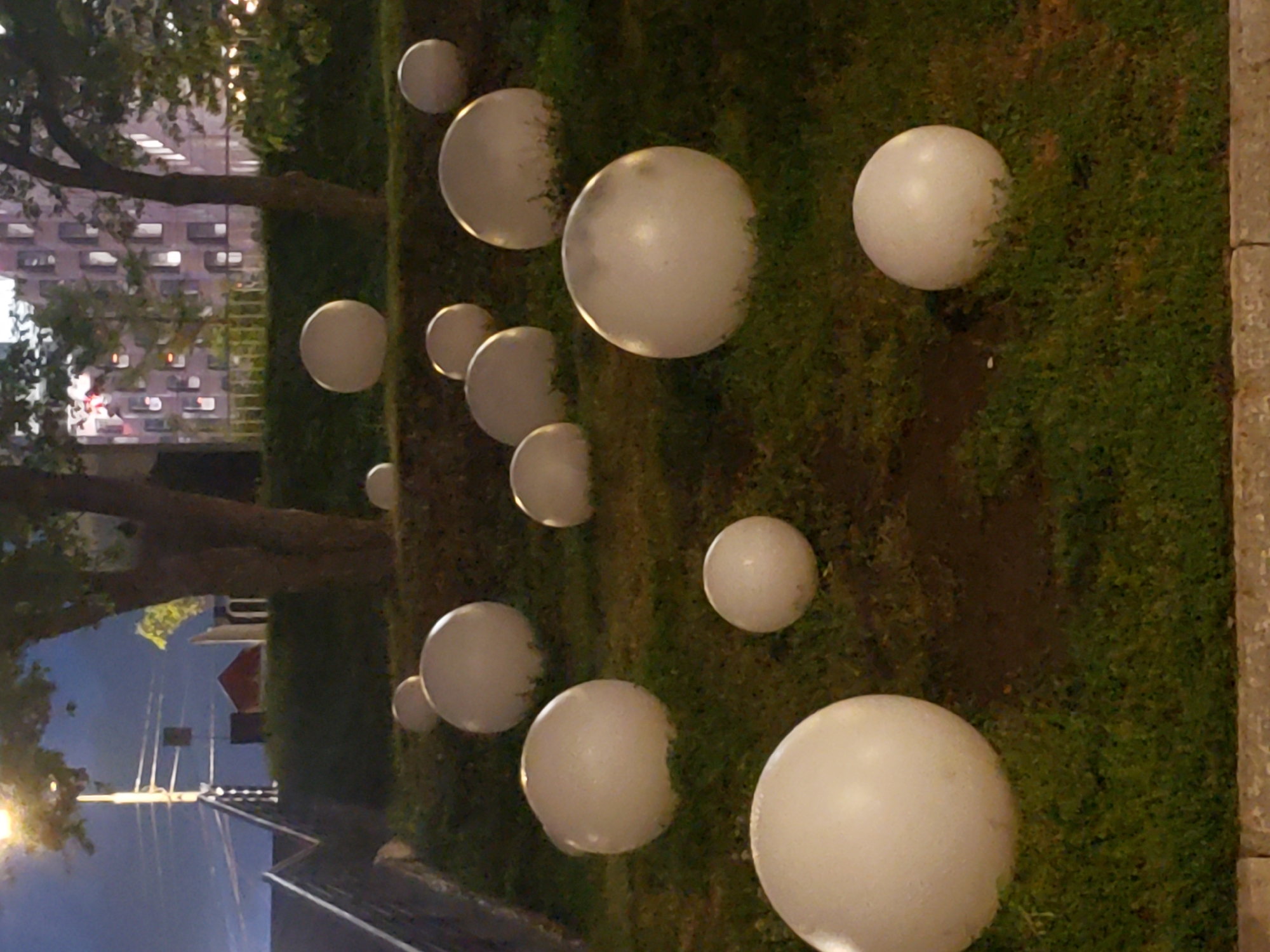 The image captures an outdoor scene with multiple illuminated white spheres scattered across a grassy area. The spheres vary in size and appear to be spherical lights, possibly garden decorations or art installations. They are lit from within, casting a soft glow on the surrounding grass. Some of the spheres are partially obscured by shadows or the terrain, suggesting an uneven ground.
In the background, a building with many windows is visible, hinting at an urban or suburban setting. Trees with dark branches are also present, suggesting it might be nighttime or dusk. The lighting from the spheres and potentially streetlights creates a somewhat ethereal or magical atmosphere. The overall impression is one of a decorative display set up in a park or garden area. There is no text visible in the image, nor any people. The time of day appears to be evening or night due to the artificial illumination of the spheres and the darkness of the surroundings.