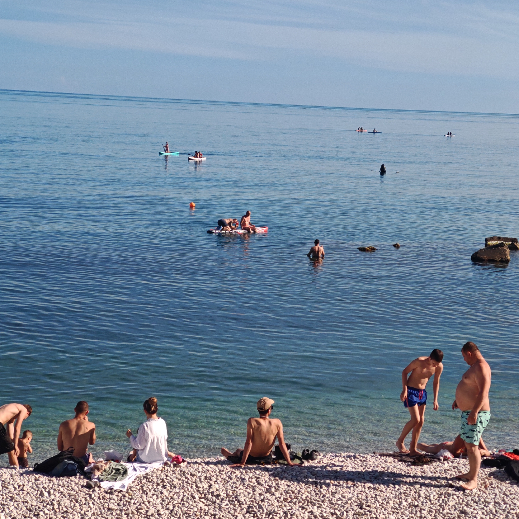 A bright, sunny day at a pebbled beach by the clear blue sea. Several people are enjoying the water and shore. In the water, two people are on a paddleboard, one sitting and one kneeling. Further out, a lone person stands in the water. On the beach, a young child stands in shallow water while a man in black shorts bends over them. Nearby, a man and woman sit on a white blanket, facing the sea. Another person sits on the pebbled shore, facing away from the camera. In the foreground, two men stand by the water's edge. The sea also contains a few small rocks and an orange buoy. The sky is a clear, pale blue with faint white clouds on the horizon.