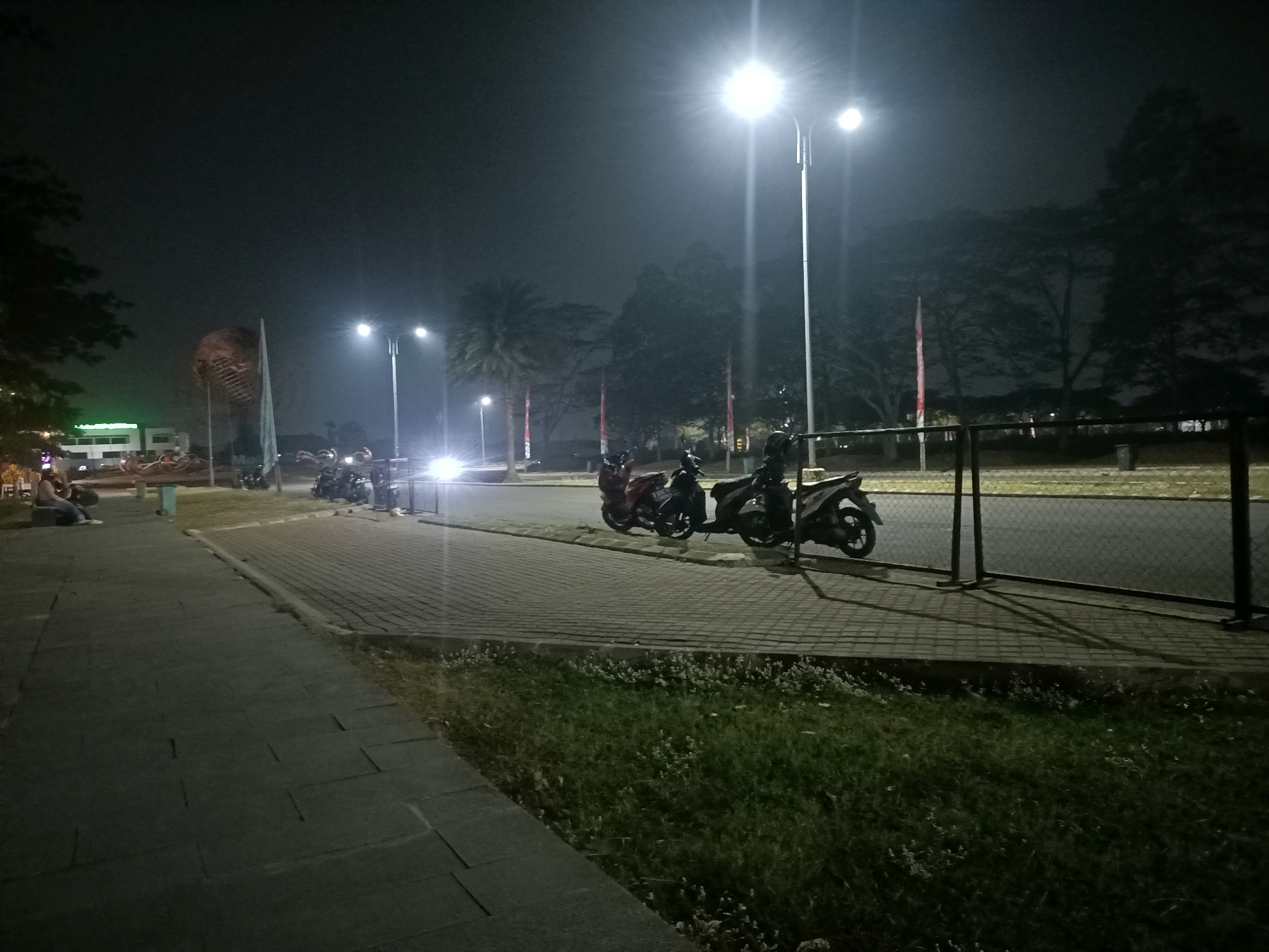 A nighttime exterior scene in Telagamurni, Indonesia, depicts a well-lit street with a paved walkway on the left and a grassy area bordering a road on the right. Several motorcycles are parked along the roadside. Two individuals are seated on a bench on the left side of the walkway, and another person stands further down. Tall streetlights illuminate the area, casting long shadows. In the background, trees and buildings are visible, along with a large, abstract sculpture. The sky is dark, suggesting it is late at night.