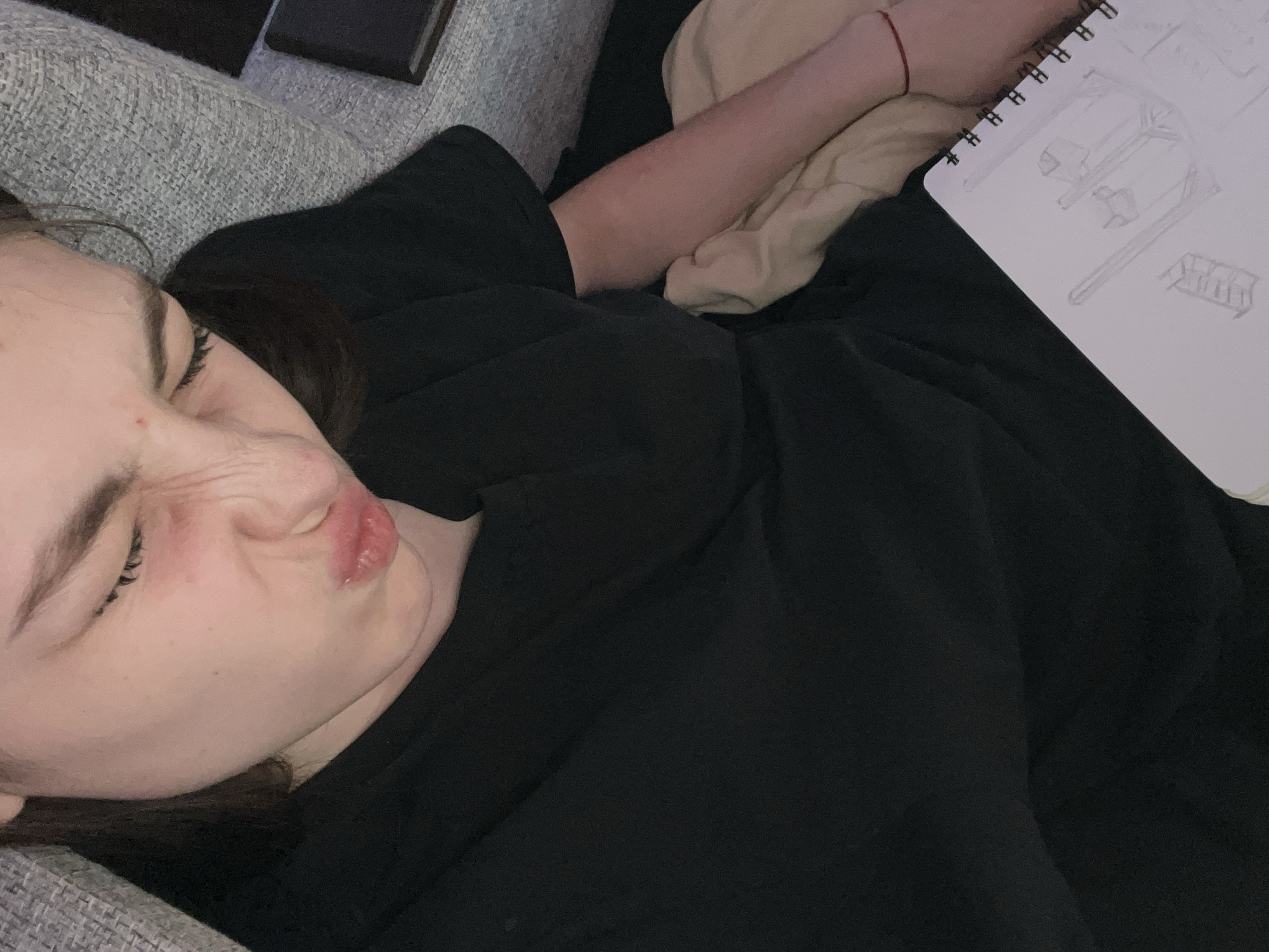 A young woman is lying on a grey couch or bed, wearing a black shirt. She is turned to her left, with her eyes closed and her face in a pucker. Next to her is a sketchbook with a drawing on it. Her left arm is on a light beige surface, possibly a pillow or blanket. There is a red string bracelet on her wrist. A black object is visible on the couch near her head. There is no evidence indicating the location is in Saint Petersburg, Russia.