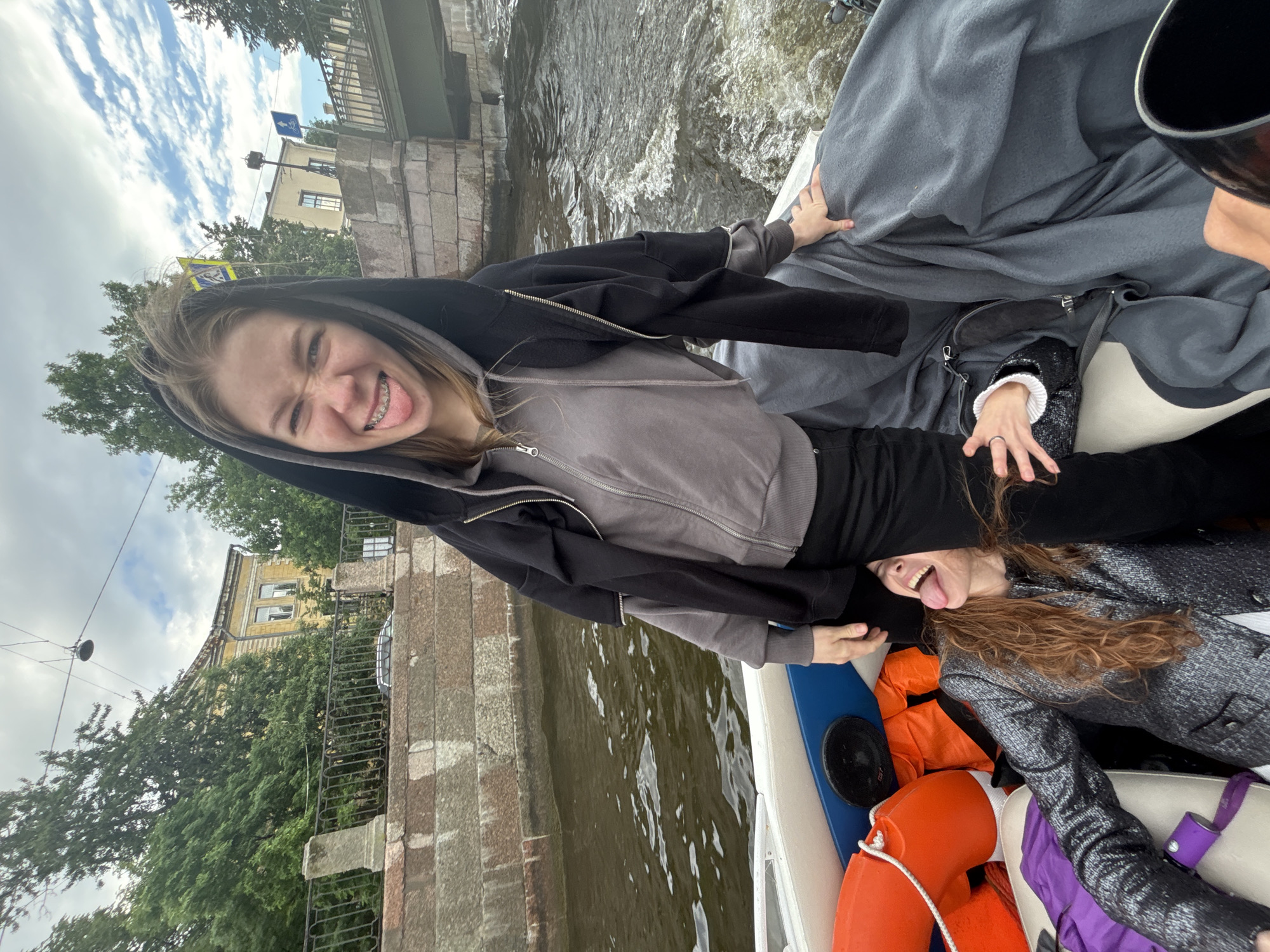 Two young women are on a boat in a canal. The woman in the foreground is wearing a black hooded jacket and a grey zip-up hoodie, with her tongue sticking out and teeth in braces visible. The woman behind her, partially obscured, is wearing a shimmering grey jacket and has her tongue sticking out. They are both in a boat with orange life preservers. The canal is lined with stone walls and trees, with buildings visible in the background. The sky is cloudy. The scene appears to be in Saint Petersburg, Russia, given the architectural style of the buildings and the presence of canals. The mood seems playful and lighthearted.