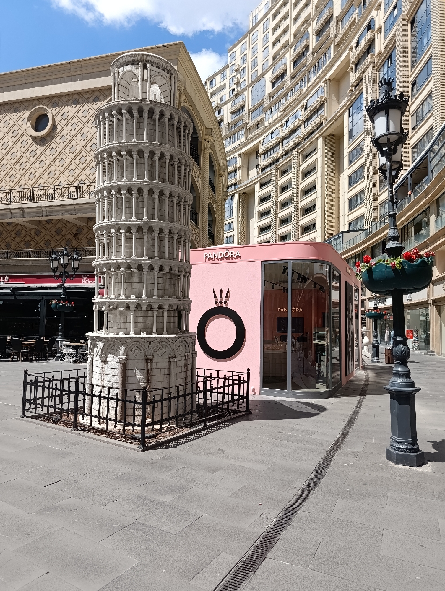 The image depicts an outdoor scene in the city of Gaziosmanpaşa, Turkey, featuring a miniature replica of the Leaning Tower of Pisa. The tower, constructed of white stone and surrounded by a black metal fence, stands prominently in the foreground. Adjacent to the tower is a modern, pink, glass-enclosed kiosk with the "PANDORA" logo displayed on its exterior. The kiosk appears to be a retail establishment. In the background, a tall, multi-story building with a beige facade and numerous windows dominates the skyline. Several streetlights, one with a planter containing red flowers, are visible along the paved walkway. The ground is paved with large grey stones. The sky above is blue with scattered white clouds, indicating daytime. No people are clearly visible in the scene.