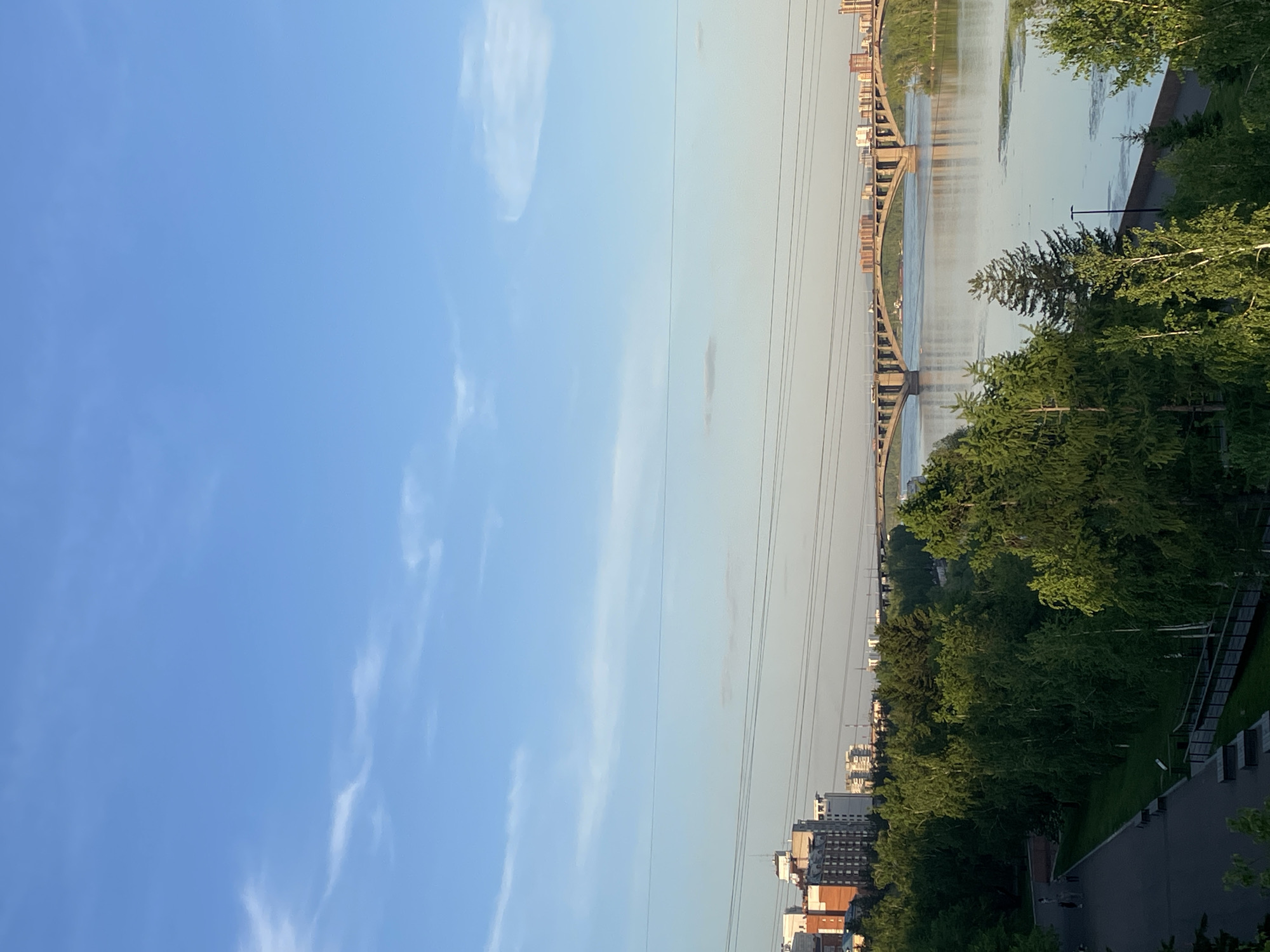 The image captures a panoramic view of a city landscape featuring a river, a prominent bridge, and a mix of urban and natural elements, under a bright, clear sky.
In the upper part of the frame, a vast expanse of light blue sky is visible, dotted with wispy white clouds. Below the sky, a wide body of water, likely a river, flows horizontally, reflecting the sky's light. Spanning this river is a large, arched bridge with multiple concrete supports, connecting the two opposing banks. Utility lines are stretched across the sky, running above and parallel to the bridge.
On the far bank of the river, to the left, a dense cluster of buildings forms a cityscape along the waterline. On the near bank, to the right and in the foreground, a vibrant green area dominated by numerous trees suggests a park or a forested river promenade. Below the treeline, a paved pathway is visible, where several small figures appear to be walking. Further into the foreground and behind the trees, more buildings are discernible, indicating urban development on this side of the river as well. A single lamppost stands tall among the trees.
The scene is bathed in clear, soft sunlight, suggesting either early morning or late afternoon. The weather appears pleasant and clear. The overall impression is one of urban tranquility, blending natural green spaces with city architecture and infrastructure. The setting aligns with a large city, such as Krasnoyarsk, Russia, which is known for its bridges over the Yenisei River. No visible text is present in the image.