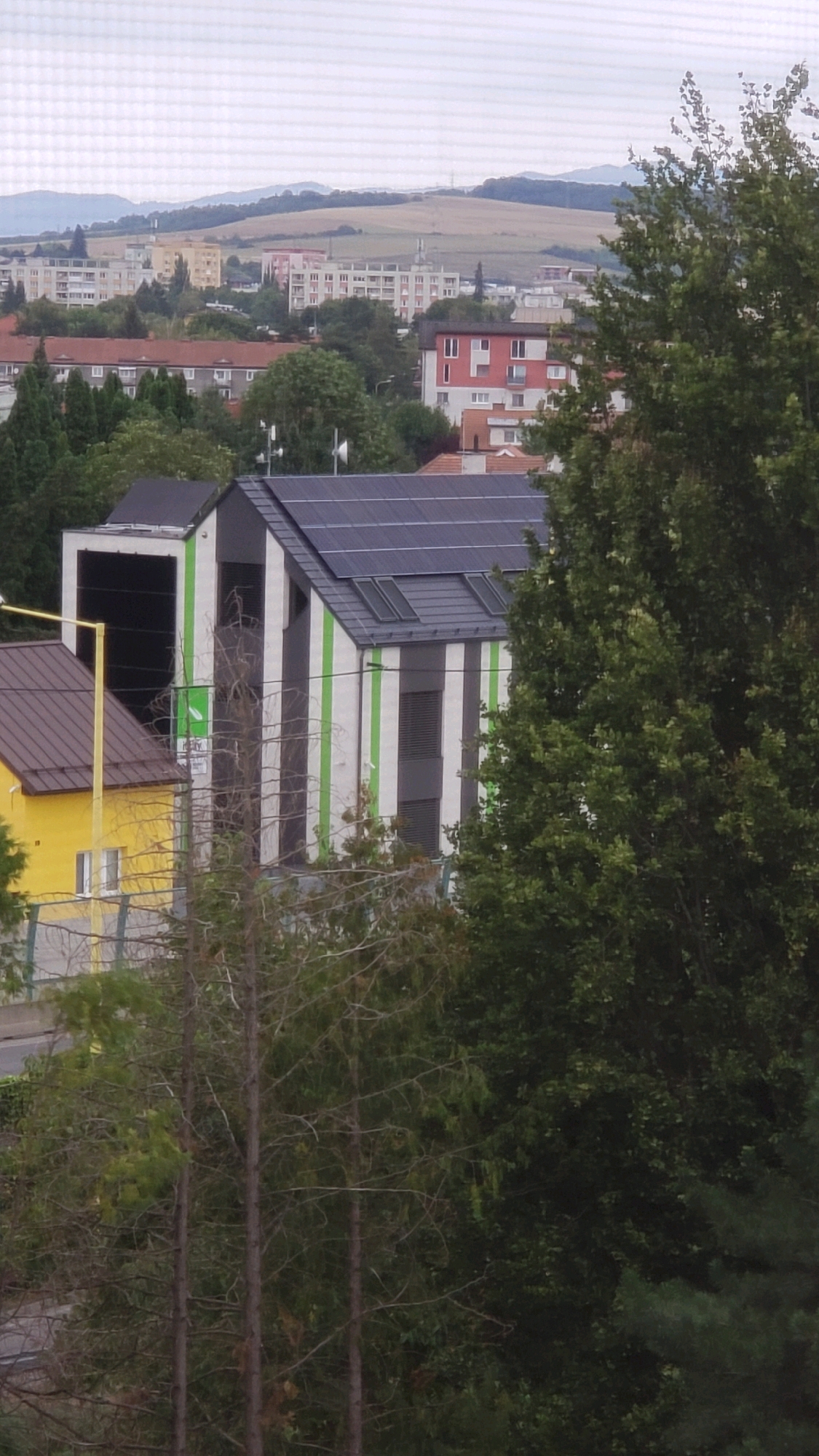 The image shows a modern building with a striking facade of vertical black and green stripes, covered with solar panels on its sloped roof. Adjacent to this building is a yellow house with a brown roof. In the background, a cityscape with apartment buildings, houses, and rolling hills under a cloudy sky can be seen. The overall scene appears to be a residential area within the city of Prešov, Slovakia. The foreground is dominated by trees, some of which are bare, indicating a possible autumn or winter season.