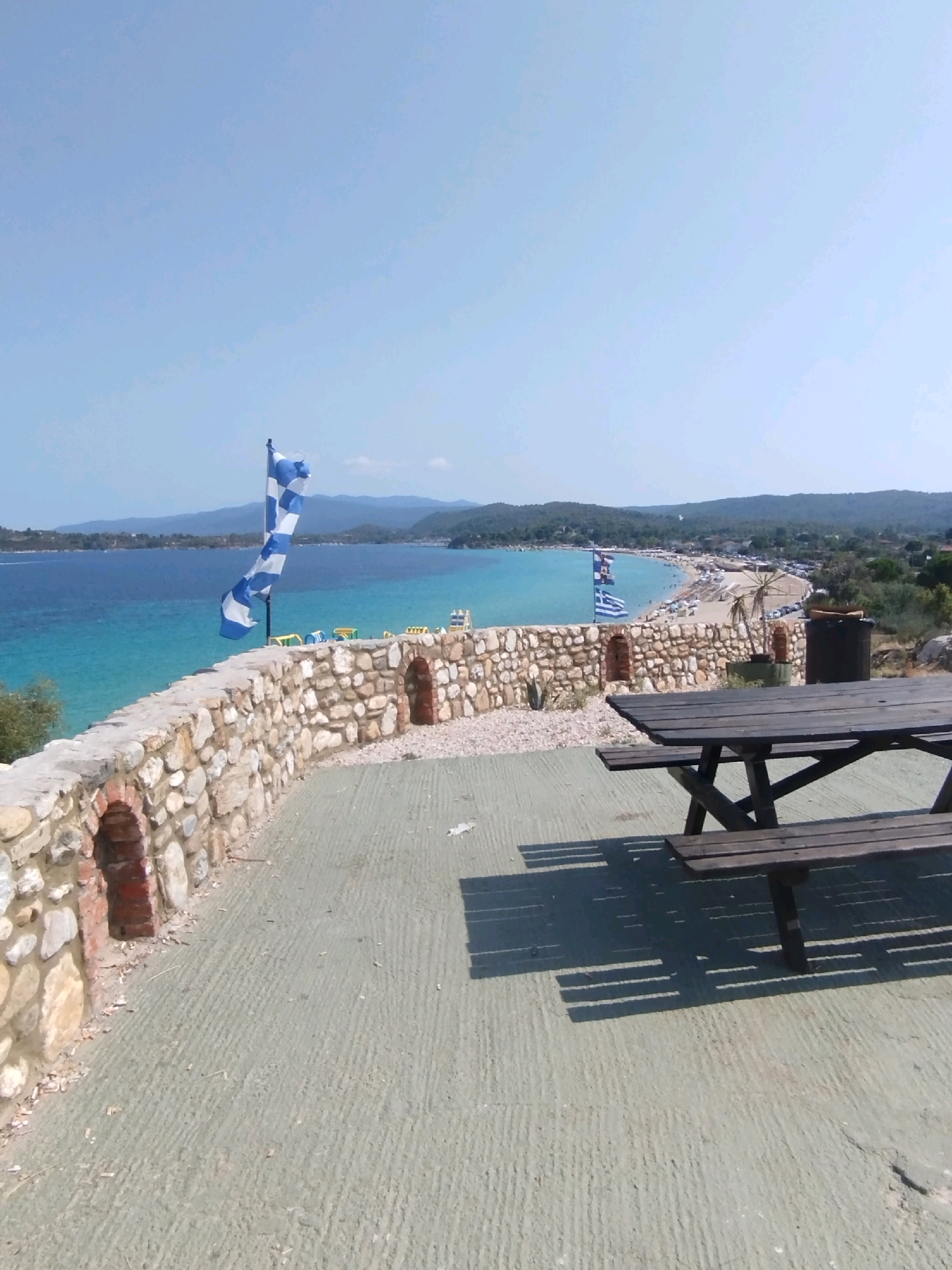 The image depicts a scenic view from a stone wall overlooking a coastal area. In the foreground, a wooden picnic table and benches are positioned on a concrete surface. Beyond the wall, the turquoise waters of a bay stretch towards a sandy beach lined with trees and buildings. A Greek flag and another checkered flag are visible on poles along the wall. Hills form the backdrop, under a clear blue sky. The scene suggests a recreational area, possibly a viewpoint or a restaurant terrace, in Sithonia Municipal Unit, Greece. There are no people or animals clearly visible in the immediate foreground or midground, though the beach appears to have some activity. The time of day is daytime, indicated by the bright sunlight and the shadows cast by the picnic table.
