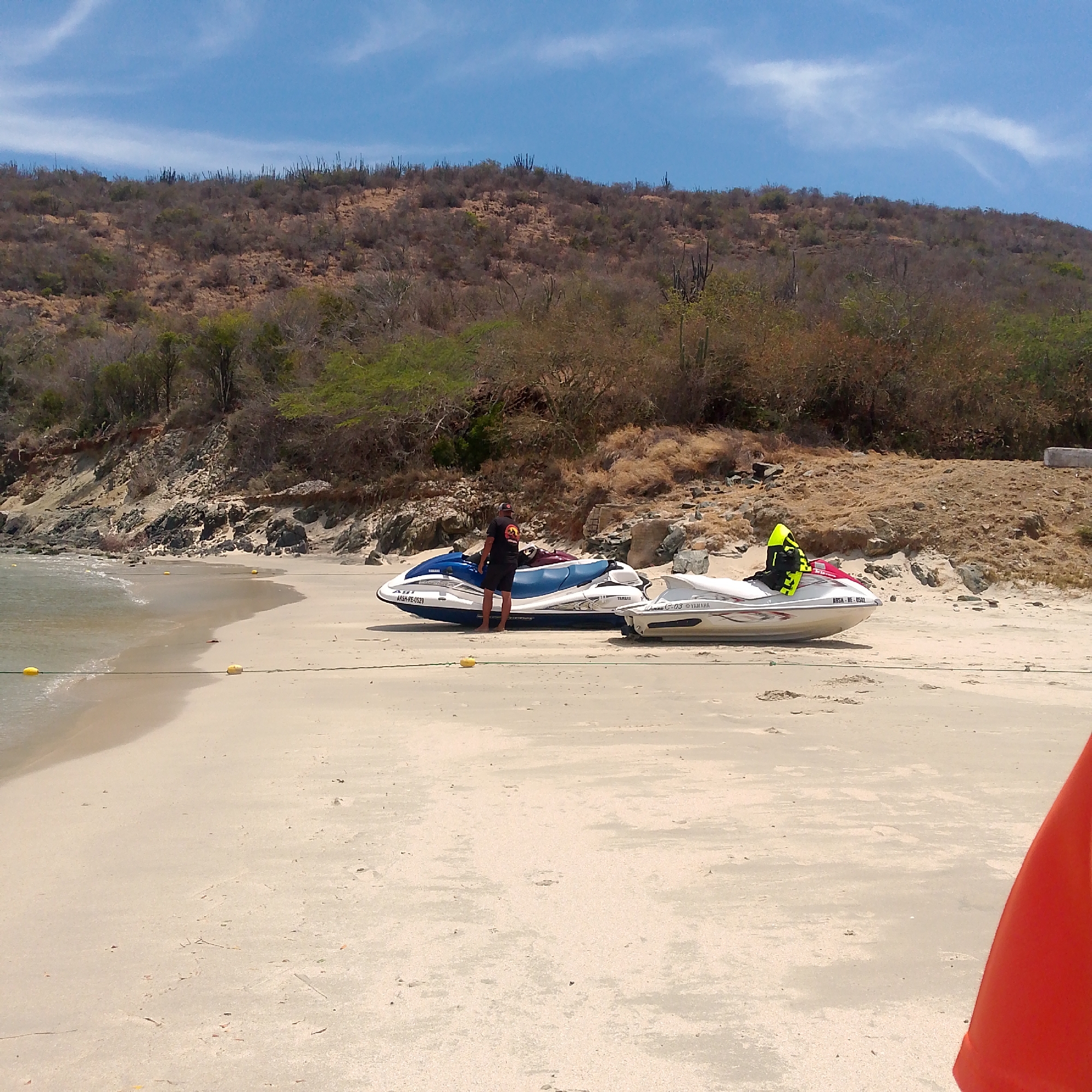 This media file captures a sunny beach scene in Pedro González, Venezuela. Two personal watercraft (jet skis) are parked on the light-colored sandy beach near the water's edge. The closest jet ski is blue and white, while the one behind it is white with red details.
A person, barefoot and wearing a black t-shirt and dark shorts, stands beside the blue and white jet ski. Another individual, clad in a bright yellow-green life vest, is visible on the white jet ski. Yellow buoys connected by a rope stretch across the sand and into the calm, clear water, possibly indicating a boundary or mooring area. An orange object is partially visible in the bottom right corner of the frame.
The background features a dry, rugged hill covered with sparse, scrubby vegetation and distinct cacti, characteristic of an arid coastal region. A rocky shoreline separates the hill from the sea. The sky is a clear, bright blue with a few wispy white clouds, suggesting a sunny day, likely midday or early afternoon.
Visible text on the blue jet ski reads "ARSH-RE-0529" and "YAMAHA". The white jet ski displays "C-03 YAMAHA" and "ARSH-RE-0502". The scene portrays a recreational setting centered around watersports.
