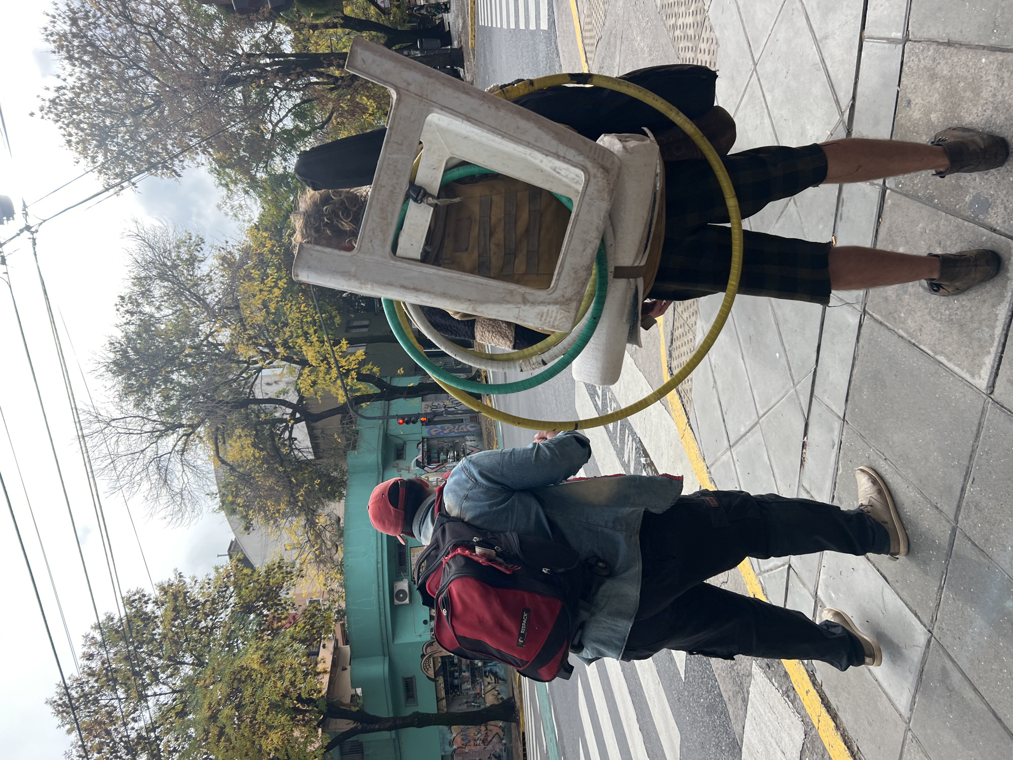 Two pedestrians stand on a sidewalk next to a crosswalk in Buenos Aires, Argentina. One pedestrian wears a red baseball cap, denim jacket, black pants, and tan shoes. They have a red backpack and are facing forward. The other pedestrian, seen from behind, carries a white plastic chair, yellow and green hoops, and other items strapped to their back. They wear a black jacket, plaid shorts, and brown shoes. Trees and a building are visible in the background.