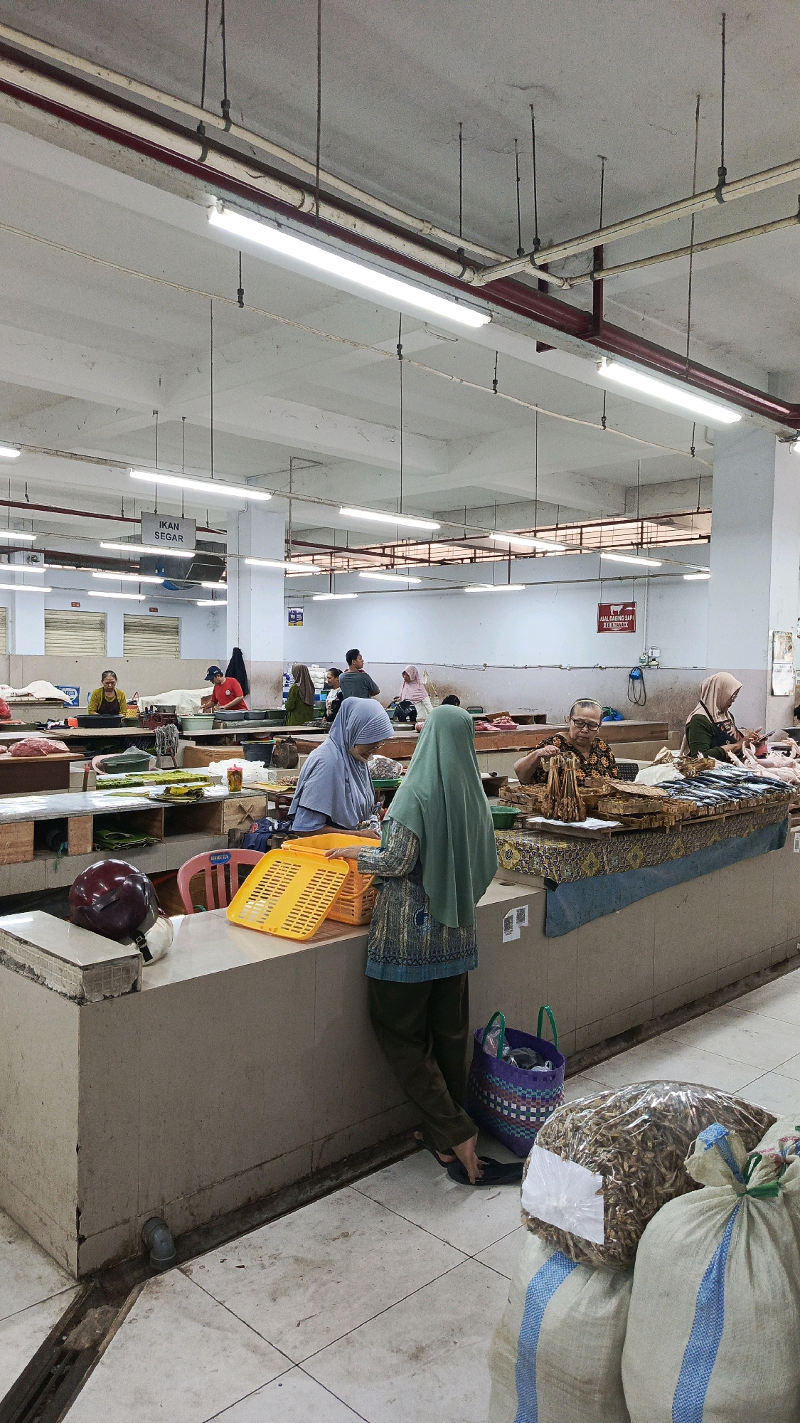 This is an indoor marketplace scene in Ponorogo, Indonesia, likely a fish market given the signage and the presence of fish on display. In the foreground, two women are engaged in a transaction. One woman, wearing a green hijab, is handing a yellow plastic basket to another woman, who wears a light blue hijab and is partially obscured. Behind them, a stall displays various dried fish and some fresh fish. Other vendors and customers are visible in the background, with stalls set up along long counters. The environment is utilitarian, with a concrete floor, tiled walls, and exposed ceiling pipes and fluorescent lighting. Bags of dried goods, possibly fish, are stacked in the lower right corner. The overall impression is of a bustling, active market.