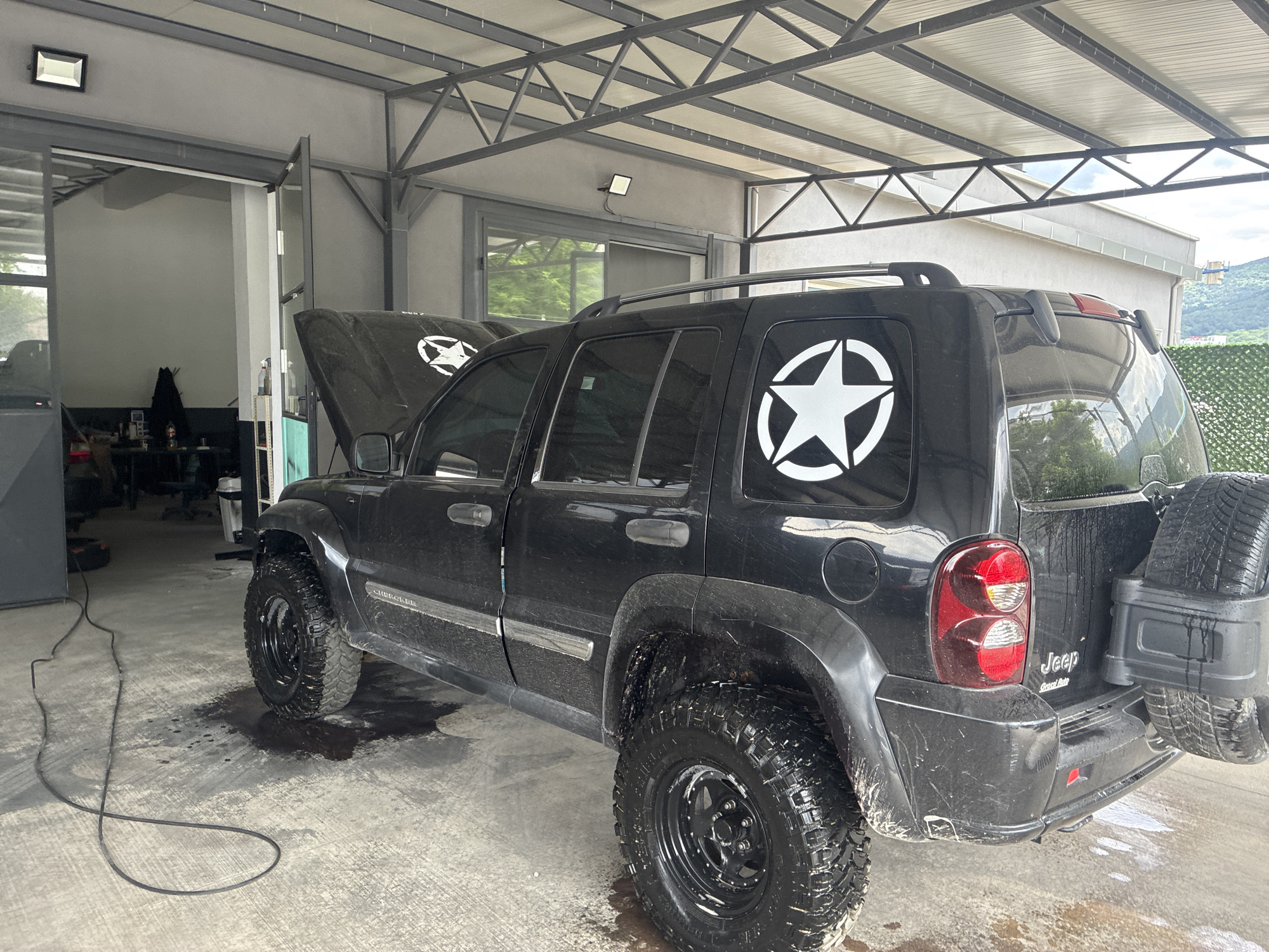 The image shows a black Jeep Liberty SUV parked indoors, likely in a garage or repair shop. The car is dirty, with mud splattered on the tires and body. The hood is open, revealing a white star decal. A similar decal is visible on the rear window. The vehicle is equipped with an external spare tire carrier on the back. The garage has a concrete floor and metal support beams for the roof. There are various tools and equipment visible inside the garage. The city and state are unknown.