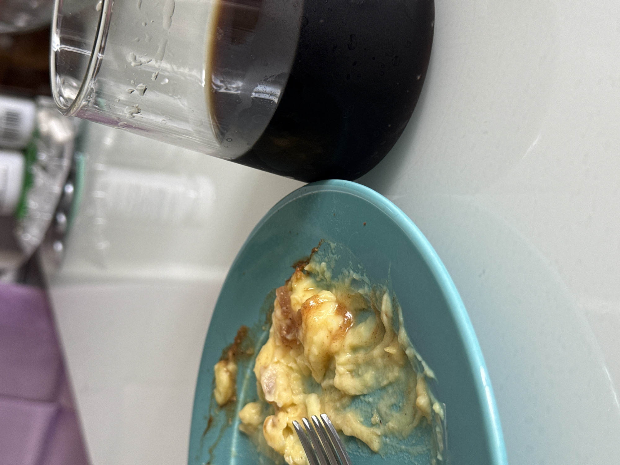 The image shows a partially consumed meal on a light blue plate with a fork. The meal appears to be scrambled eggs with some sort of topping, possibly syrup or sauce. Above the plate is a glass containing a dark liquid, likely coffee or a similar beverage. The background includes a white surface, possibly a countertop or table, and blurry elements suggesting the scene is indoors, potentially in a kitchen or dining area. The location is not specified, and is given as Unknown, Unknown.