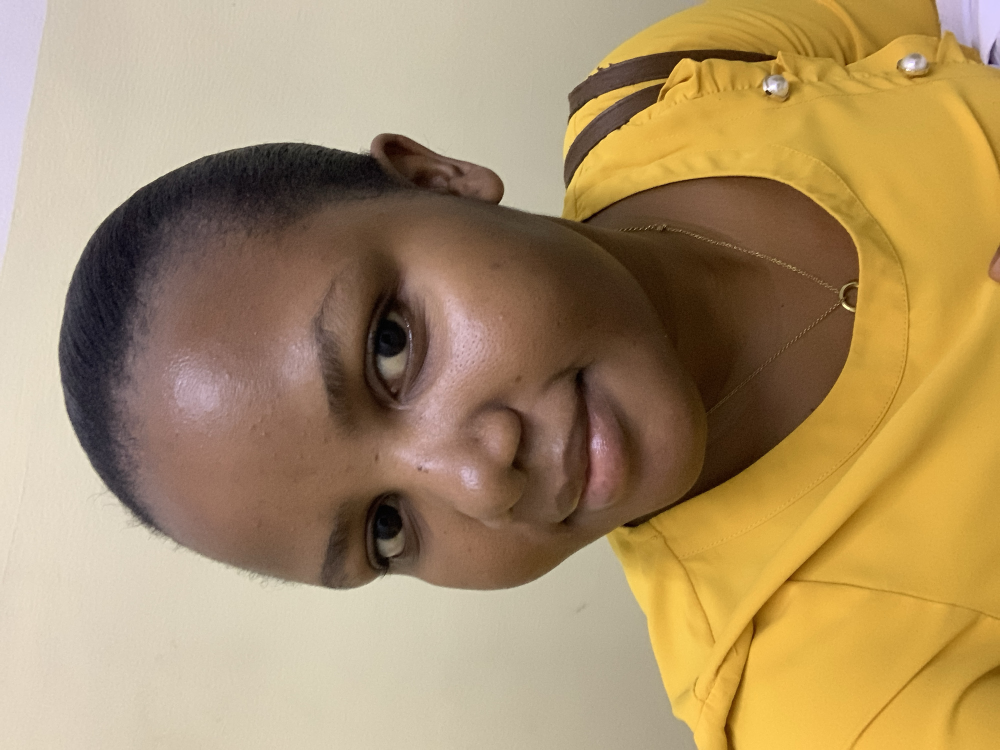 A young woman with dark skin and short hair is looking at the camera. She is wearing a yellow shirt with brown stripes and silver buttons. A gold necklace is visible around her neck. The background is a light beige or yellow wall. The image does not provide specific information to confirm the location in the city of Dar es-Salaam, Tanzania.