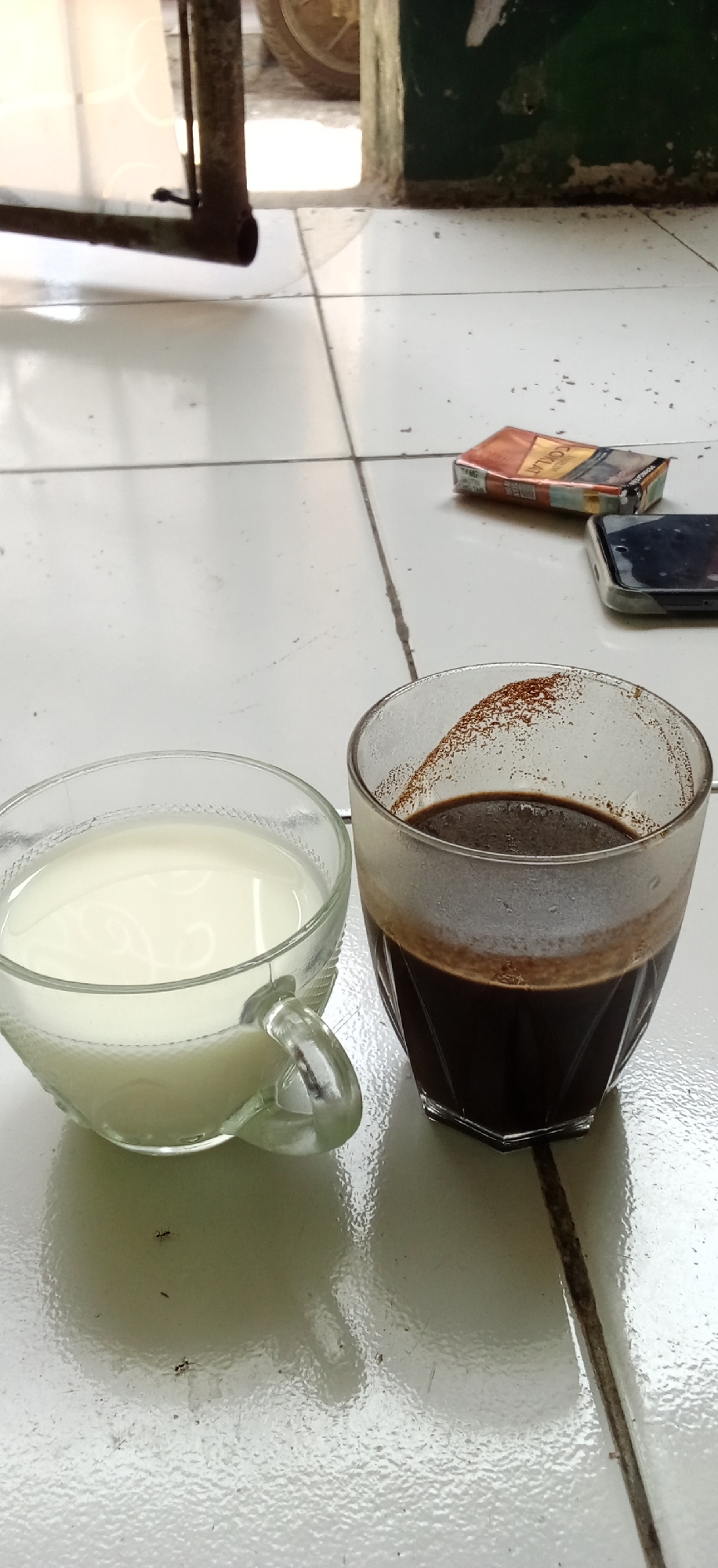 The image captures a casual daytime scene on a white tiled floor in what appears to be a residential setting in Banjaran, Indonesia. Two clear glass cups are prominently featured: one filled with white milk, and the other with dark brown coffee, showing some residue along its inner rim.
To the right of the cups, a crumpled pack of "COKLAT" brand cigarettes is visible, with "14MG" printed on it, lying next to a dark smartphone. In the background, hints of the surroundings include a rusty metal frame or leg, a segment of a dark green painted wall, and part of a wheel, suggesting a semi-outdoor area or a modest interior space. The bright, natural illumination indicates it is daytime. Several small ants are seen crawling on the white tiles near the milk cup. The overall scene conveys a simple, relaxed moment, possibly a coffee and milk break.