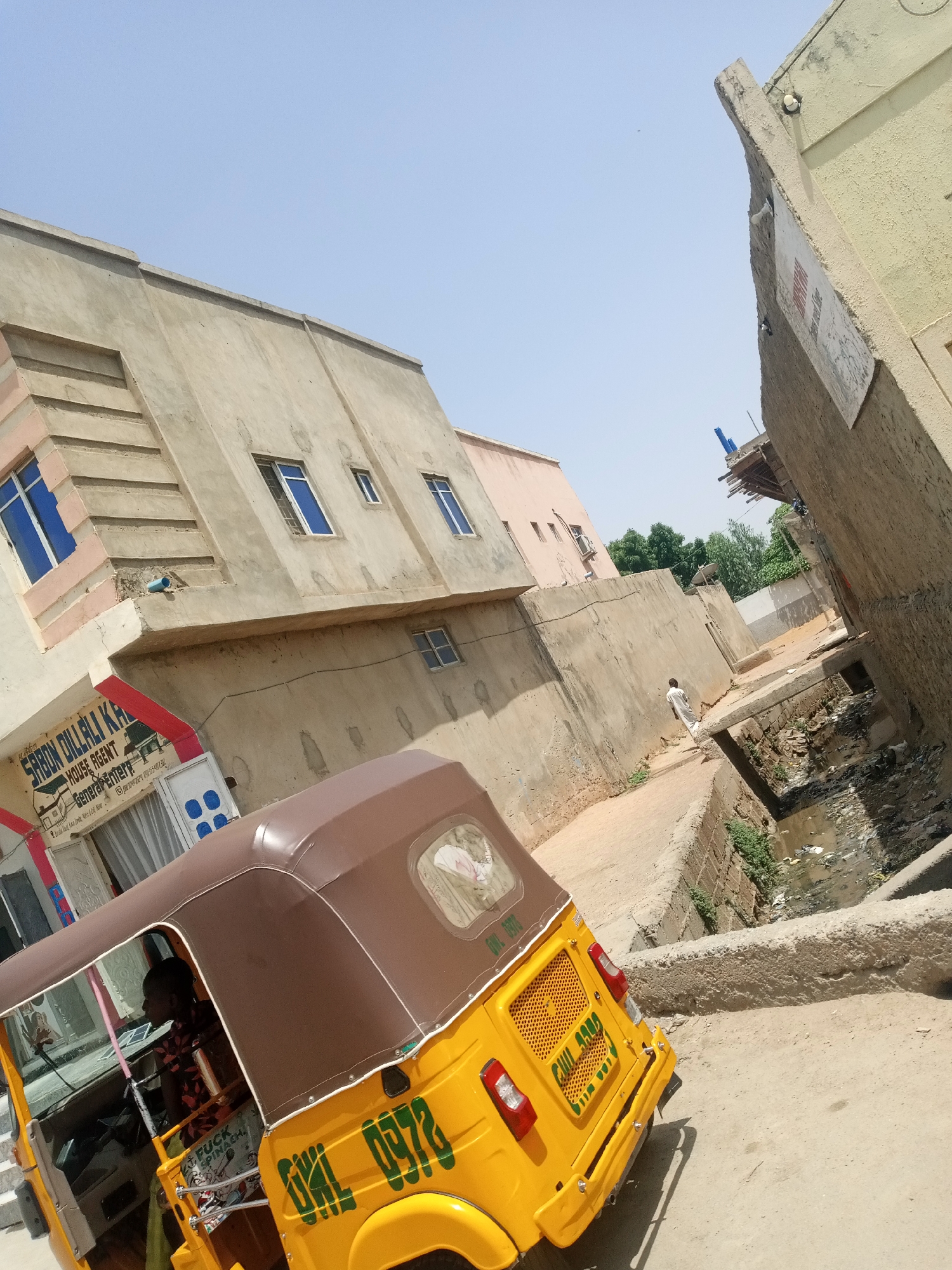 In a narrow urban alleyway, a yellow auto-rickshaw is parked on a dusty dirt road. The vehicle's brown canopy is visible, and the side displays green lettering with the number "0978" and other unreadable characters. A partially obscured person is seated in the driver's seat. To the left of the auto-rickshaw, a building displays a sign with red and white lettering that reads "SABON DILLALI KA HOUSE AGENT". Further down the alley, a person dressed in a light-colored garment walks away from the camera. The alley is flanked by concrete buildings, some appearing weathered, and a ditch filled with debris and murky water runs alongside the path. The sky is clear and bright blue.
