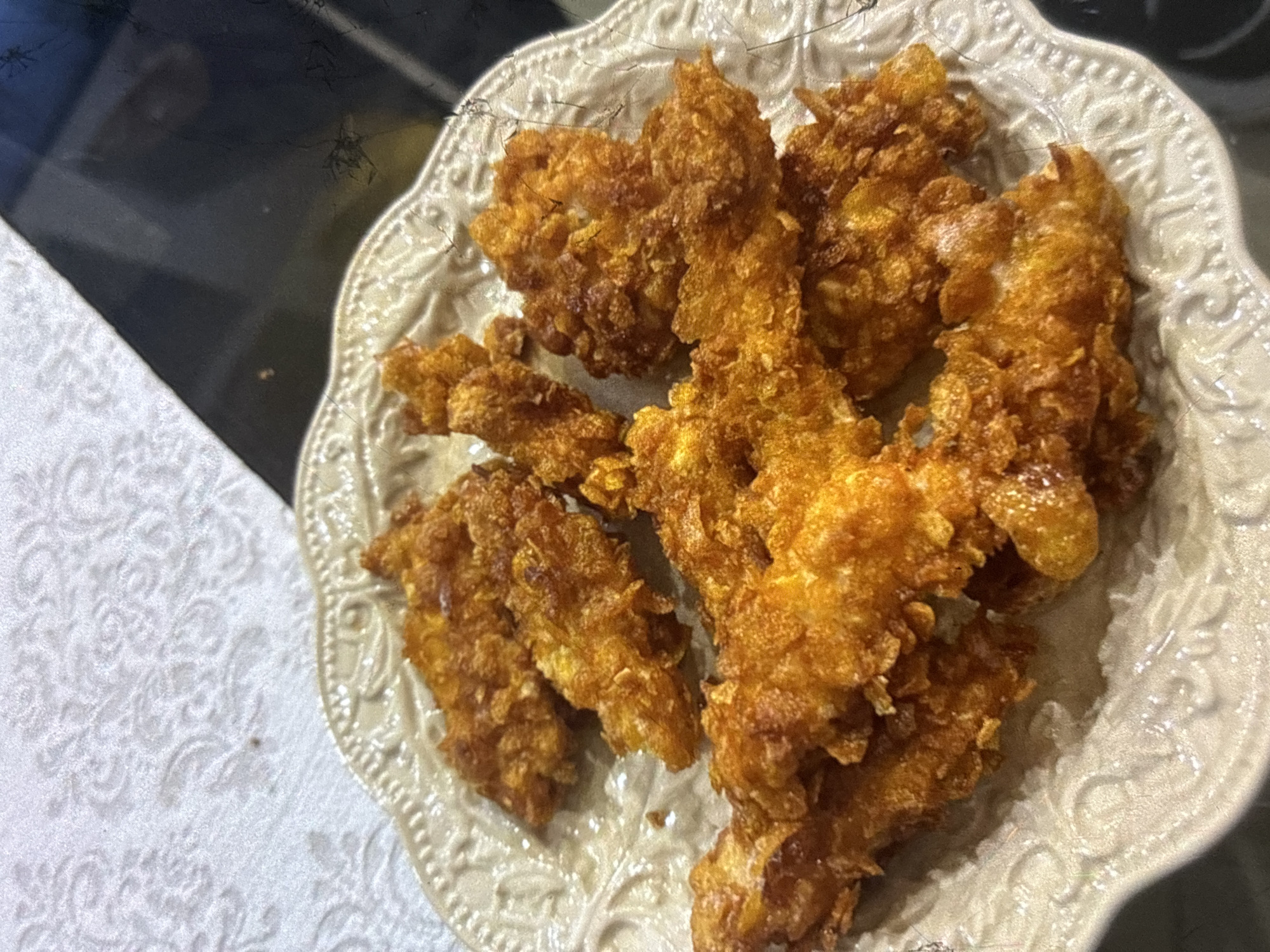 A close-up, overhead shot shows a pile of golden-brown, breaded chicken tenders arranged on a decorative, cream-colored plate. The plate has a raised, intricate pattern around its rim. The chicken tenders appear crispy and are clustered together, obscuring much of the plate. The background consists of a white, textured tablecloth on the left and a dark, reflective surface on the right, possibly a table or countertop. There is no indication of the city of Orihuela, Spain.
