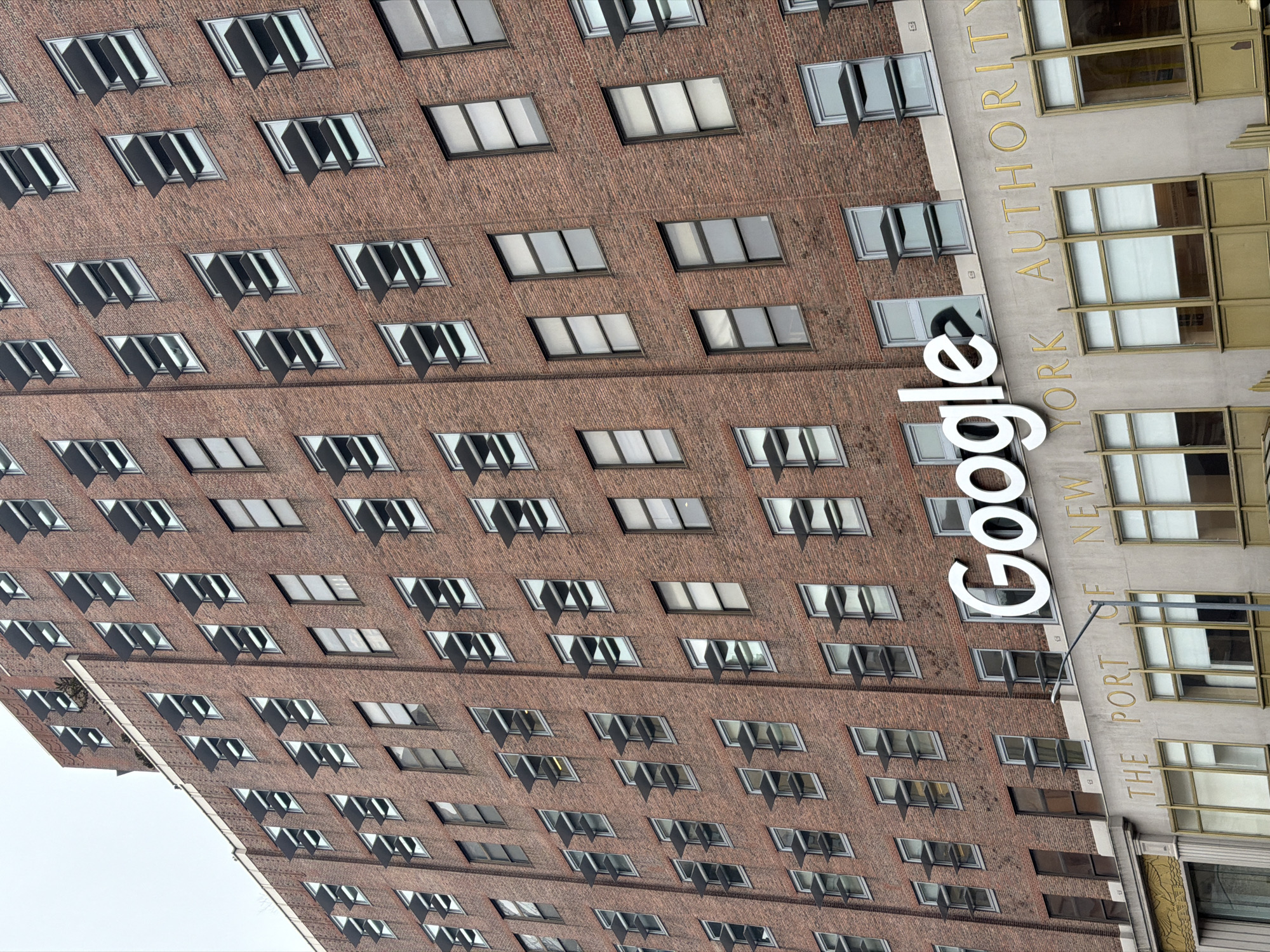 The image shows a partial exterior view of a building in New York City, United States of America. The building is brick with numerous windows, some with awnings. A large, white "Google" sign is affixed to the building's exterior. To the right of the Google sign, the phrase "The Port of New York Authority" is visible on the adjacent section of the building. The photograph is taken at an angle, with the building appearing to stretch upwards.
