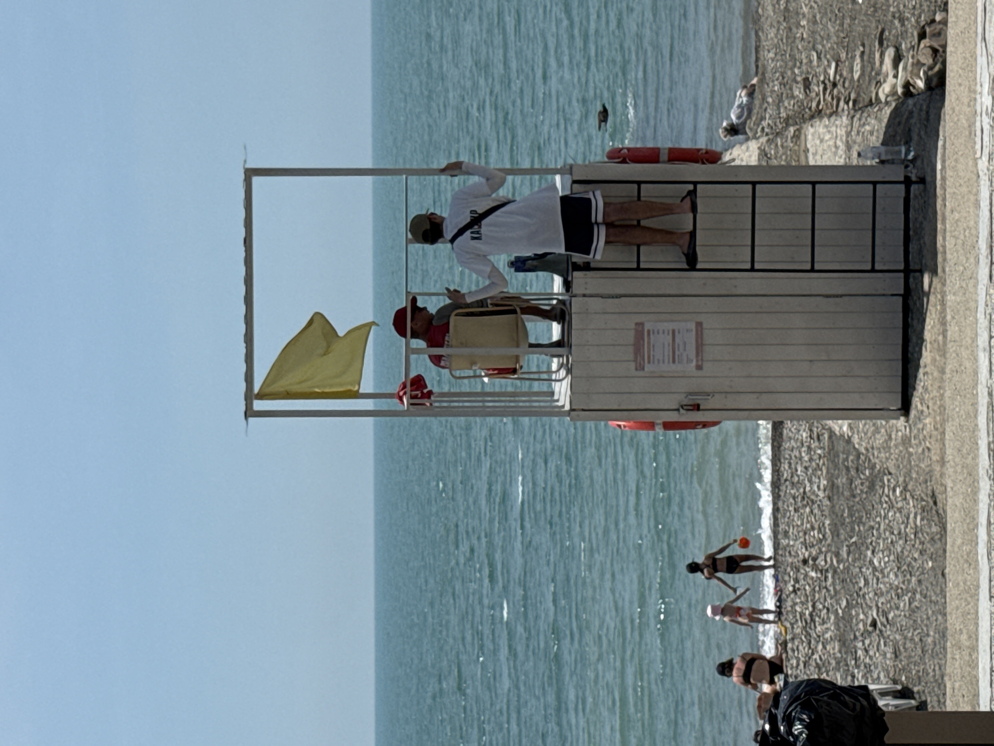 This image depicts a sunny daytime scene on a pebble beach in Sochi, Russia, alongside a calm, blue-green sea under a clear sky.
A two-story lifeguard stand dominates the mid-ground. The lower section of the stand is constructed of light-colored vertical wood paneling, featuring two red lifebuoys mounted on its exterior. A ladder with black rungs extends from the ground to the upper platform on the right side of the stand. A rectangular sign with text is affixed to the front paneling. The upper section consists of a metal frame supporting a yellow flag, partially unfurled to the left, and a small shaded roof.
On the upper platform, two men are present. One man, wearing a red cap and dark shirt, is seated in a light-colored chair on the left, facing right. The second man stands on the right, with his back to the viewer, looking out towards the sea. He wears a white long-sleeved shirt with the word "KA(or R)P" visible on the back, dark shorts, a baseball cap, and dark flip-flops, and is holding onto the metal frame.
Several individuals are on the pebble beach and at the water's edge to the left of the lifeguard stand. An adult in a dark swimsuit is seated on the pebbles. Adjacent to them, a small child in a light-colored swimsuit and hat stands at the water's edge, potentially holding hands with another adult. Further right, an adult in a dark swimsuit stands in the shallow water, holding a red bucket. On the far right, near the water line, a person in a light-colored top is lying prone on the pebbles.
A dark, indistinct object, possibly a bird or debris, is visible floating in the sea to the right of the lifeguard stand. In the immediate foreground, a black plastic bag and a white object are partially visible on the left. A clear plastic water bottle lies on the ground near the base of the lifeguard stand on the right. A concrete or paved surface runs along the foreground, parallel to the beach.