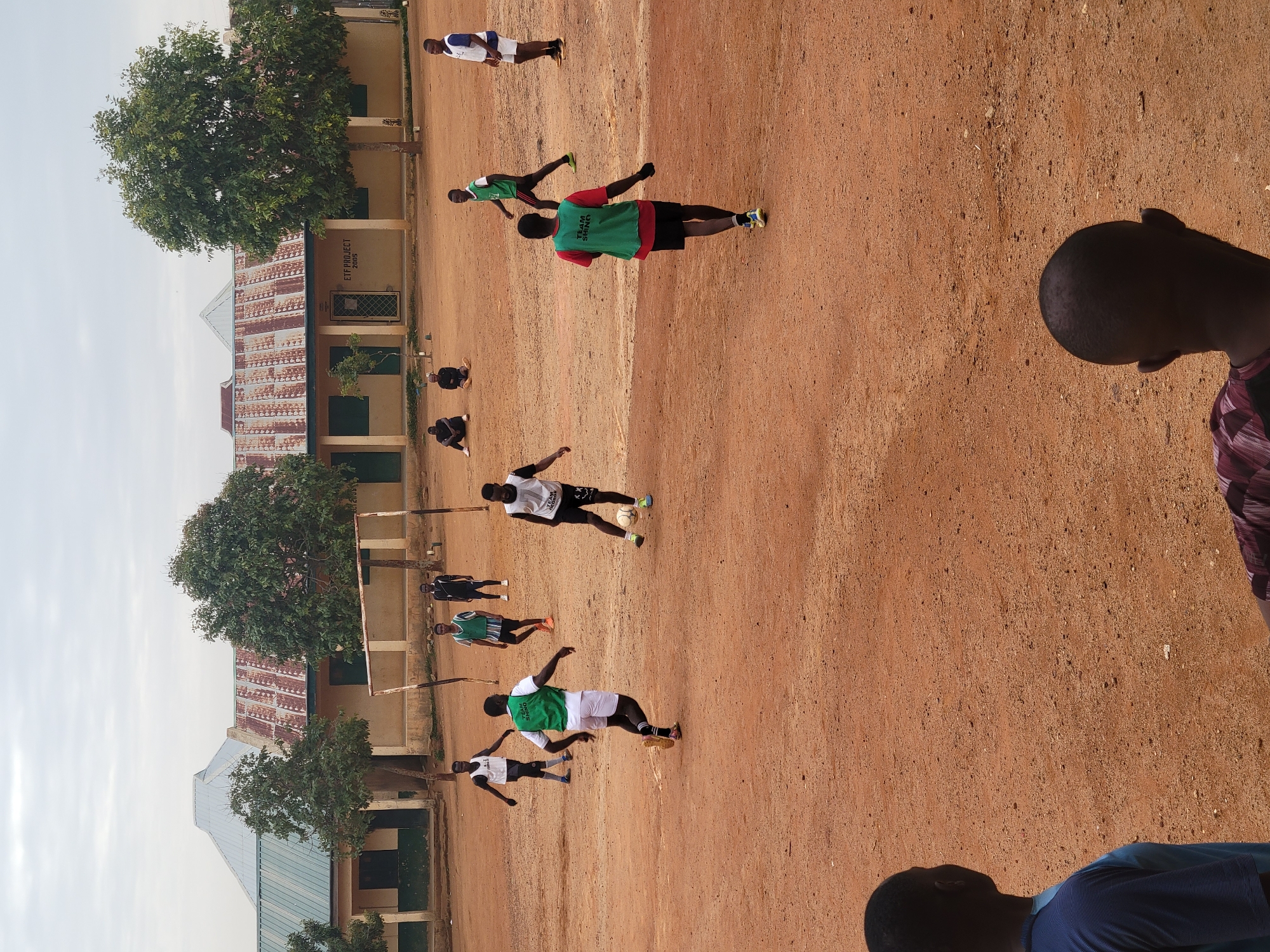 The image depicts a soccer match in progress on a red dirt field in Jos, Nigeria. Multiple players wearing different uniforms (white shirts and black shorts; green vests over red shirts; white shirts and green shorts; blue and white uniforms) are scattered across the field. Some are actively running and kicking a soccer ball. The background features a building with a corrugated metal roof and trees bordering the field. The sky is overcast. Parts of two people are visible in the foreground.
