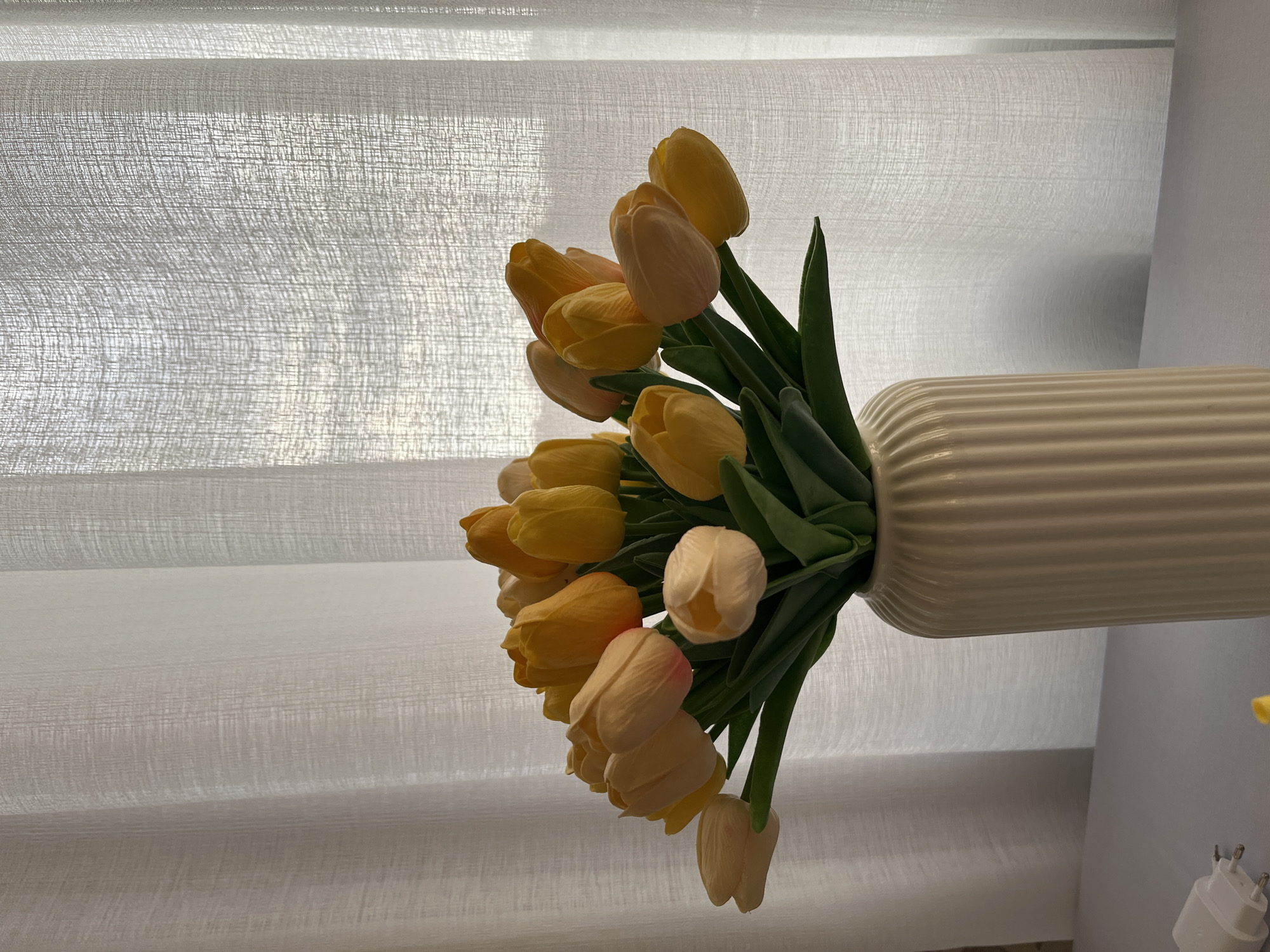 A bouquet of yellow and light pink tulips is arranged in a fluted, cream-colored vase. The vase sits on a light gray surface, and in the lower left corner, a white electrical adapter with two pins is visible. The background consists of sheer white curtains that are lit from behind, allowing some light to filter through. The overall scene is indoors and appears to be a still life composition. There are no people or animals present in the image, and no specific location in Kohtla-Järve linn, Estonia can be determined from the visual information provided.