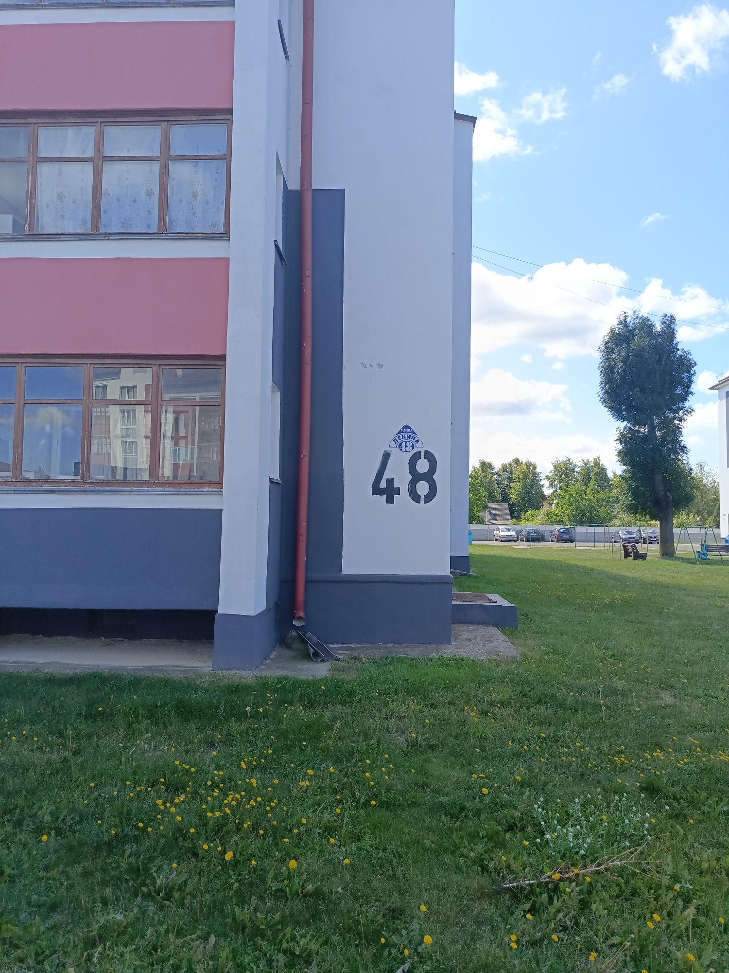 The image shows the exterior of a building with a pink and grey facade, featuring several windows. A red downspout runs vertically along the white section of the building, where the number "48" is stenciled in black. A circular emblem with Cyrillic lettering above the number indicates the location. The building is situated in a grassy area with scattered yellow wildflowers, and in the background, there are trees, cars parked along a fence, and a swing set. The sky is blue with white clouds, suggesting a daytime setting in a residential or municipal area within the city of Ivatsevichy, Belarus.