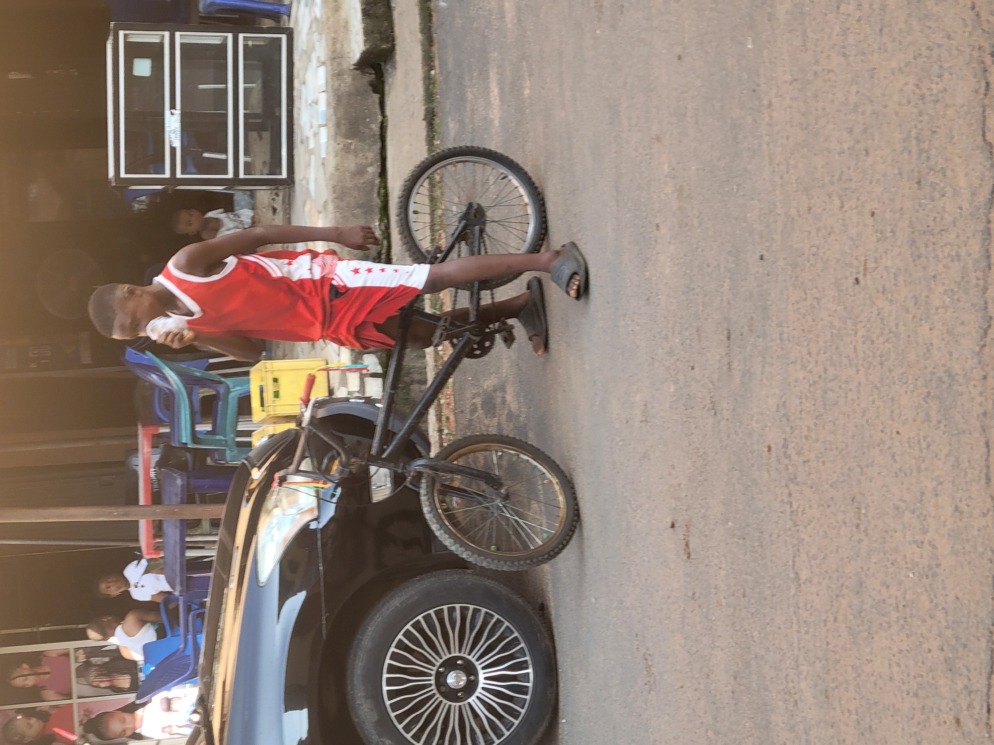 A young Black male, wearing a red sleeveless jersey with white trim and a red and white patterned fabric tied around his waist, drinks from a white cup. He is standing next to a bicycle, with the front wheel positioned in front of his right foot. The bicycle has a black frame and thin tires. Behind him, a black car is parked, with its front wheel visible. In the background, there is a building with open doorways, revealing people sitting at tables and on chairs. A tall, multi-tiered display case with glass shelves is visible to the right of the young male. The scene appears to be outdoors, possibly on a street in Onitsha, Nigeria, given the context provided. The ground is paved with asphalt.