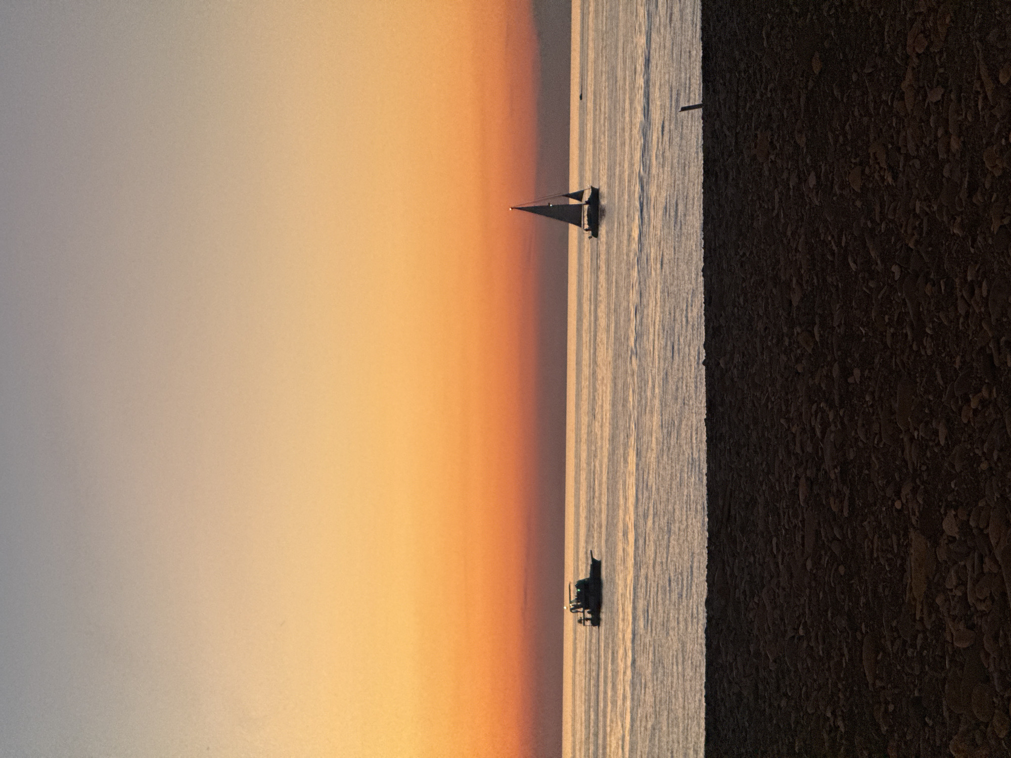 The image depicts a sunset over the Black Sea in Batumi, Georgia. The sky transitions from a pale gray at the top to a vibrant orange and then a deep red near the horizon. Two boats are visible on the water: a small fishing boat to the left and a sailboat to the right. The water is calm, reflecting the colors of the sky. In the foreground, a rocky beach is visible, with dark, rounded stones. The overall atmosphere is serene and peaceful.