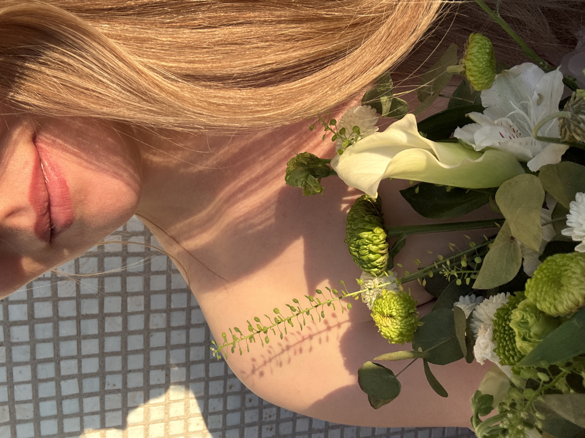 A close-up, angled shot features a woman's face, shoulder, and hair intertwined with a bouquet of flowers. The woman's face, with closed eyes and visible lips, is positioned in the upper left. Her blonde hair cascades across the top of the frame, mingling with the flowers. The bouquet, composed of white calla lilies, green chrysanthemums, eucalyptus leaves, and other greenery, rests against her shoulder. The background shows a tiled surface with geometric patterns. Light casts shadows across the woman's skin and the tiles. The location is not discernible from the image to be the city of Chaikovsky, Russia.