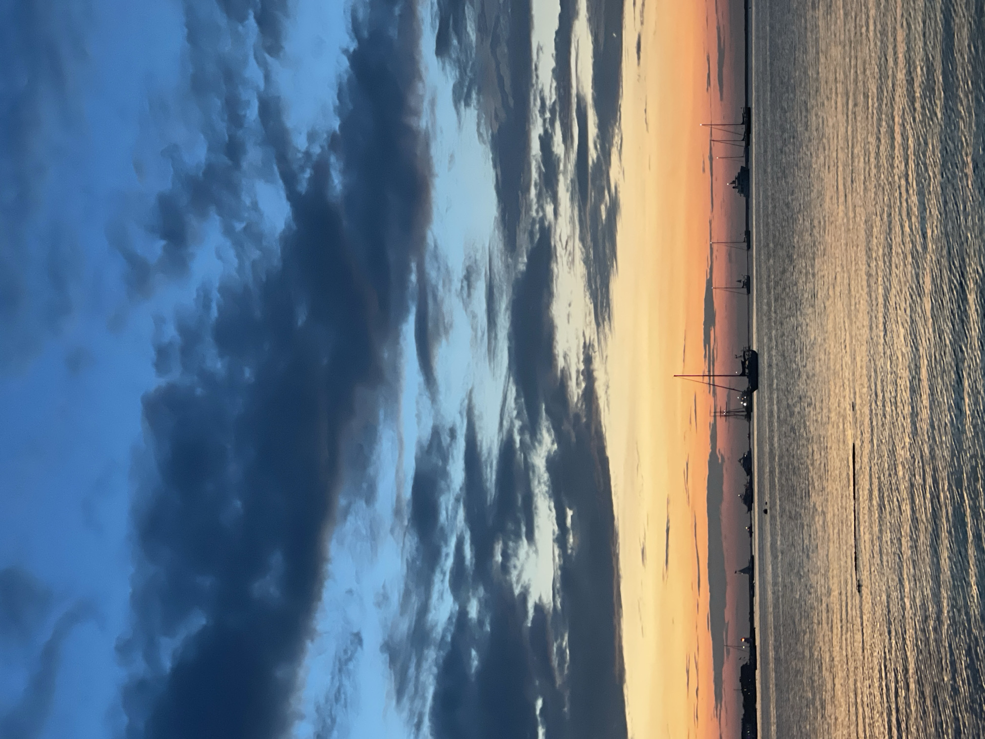 An atmospheric sunset over a body of water is captured in this vertical, full-frame photograph. The sky transitions from a deep, dark blue at the top to shades of lighter blue and then into vibrant oranges and yellows near the horizon. Dark, textured clouds are scattered across the upper half of the sky. A distant shoreline is visible with silhouetted buildings and structures, possibly a lighthouse. Several sailboats and yachts are moored on the water, their masts clearly defined against the colorful sky. The water itself reflects the colors of the sky, with rippling patterns creating texture. The scene appears to be a tranquil evening setting in the city of Nantucket, Massachusetts, United States of America, known for its picturesque harbor views.