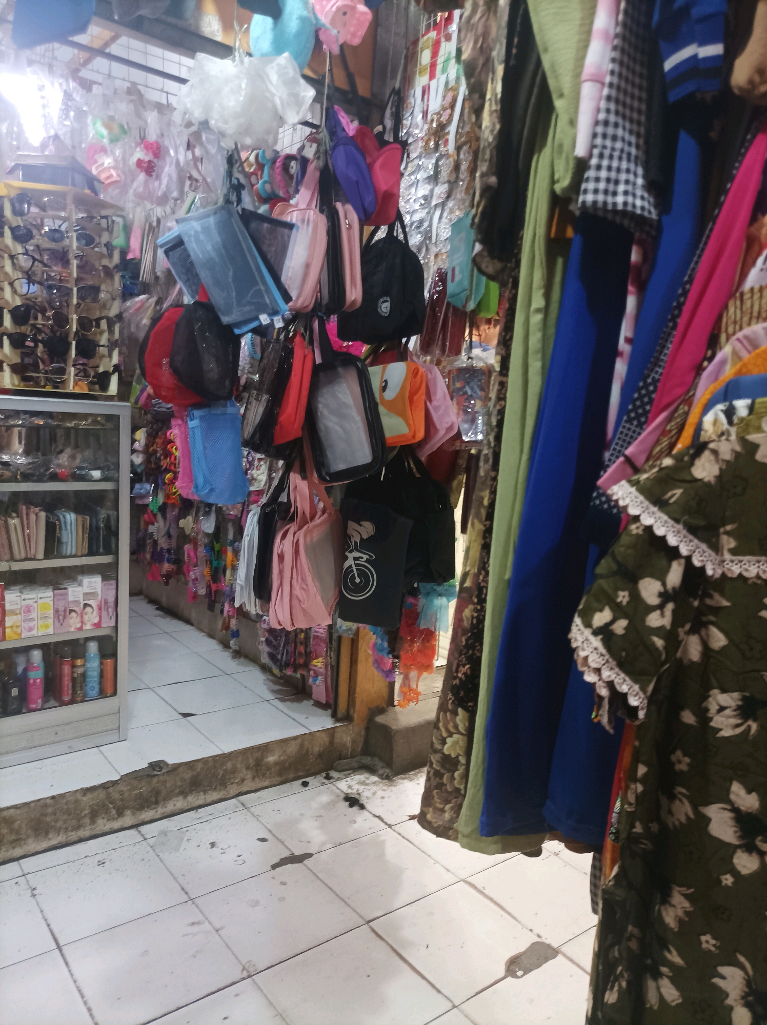 The image depicts an indoor market stall in Sumuragung, Indonesia, selling a variety of accessories and clothing items. The stall is packed with merchandise, including sunglasses displayed on a rack to the left, various pouches and bags hanging from the ceiling and a rail, and a collection of small items like keychains and hair accessories. Clothing items, such as dresses and shirts, are also visible hanging on a rail. The floor is tiled, and the overall impression is one of a bustling retail environment. The lighting appears to be artificial, typical of an indoor market.
