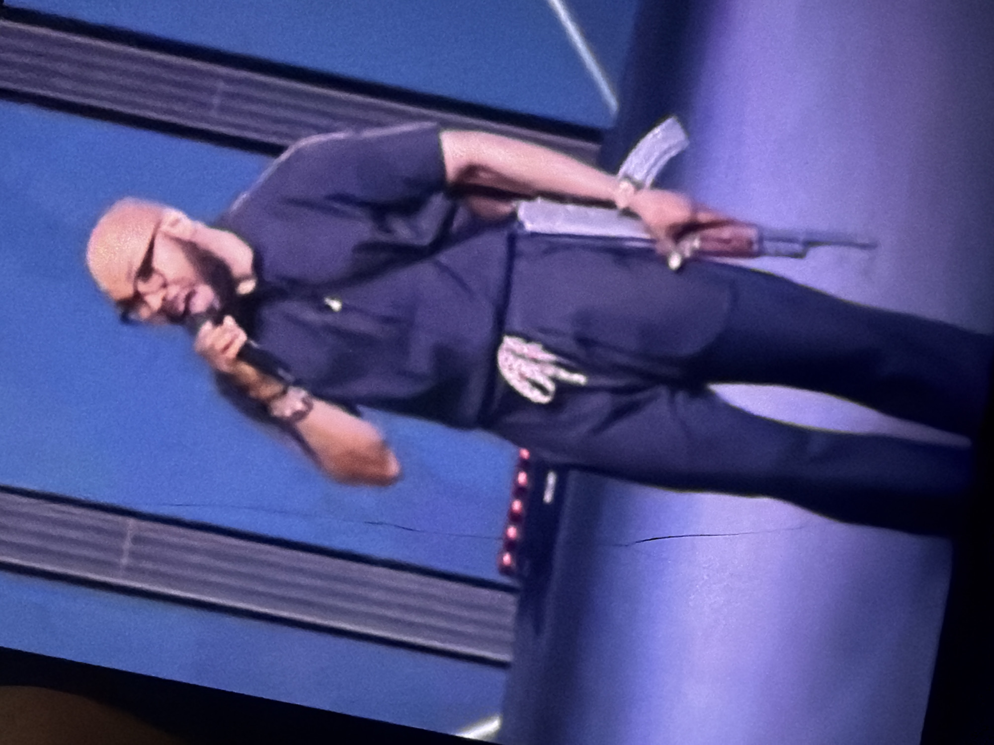 A bald Black man wearing glasses, a dark blue short-sleeved shirt, and dark blue pants is standing on a stage. He is holding a microphone in his right hand and appears to be speaking. A rifle is attached to his left hip, with the barrel pointing downwards. The man is also wearing a watch on his left wrist. The background is a blue wall with vertical ribbed panels. The stage floor is a light purple color. There are red lights in the background, below the blue wall. The image quality is poor, with visible cracks across the frame. The location of Bukuru, Nigeria is not determinable from the image.