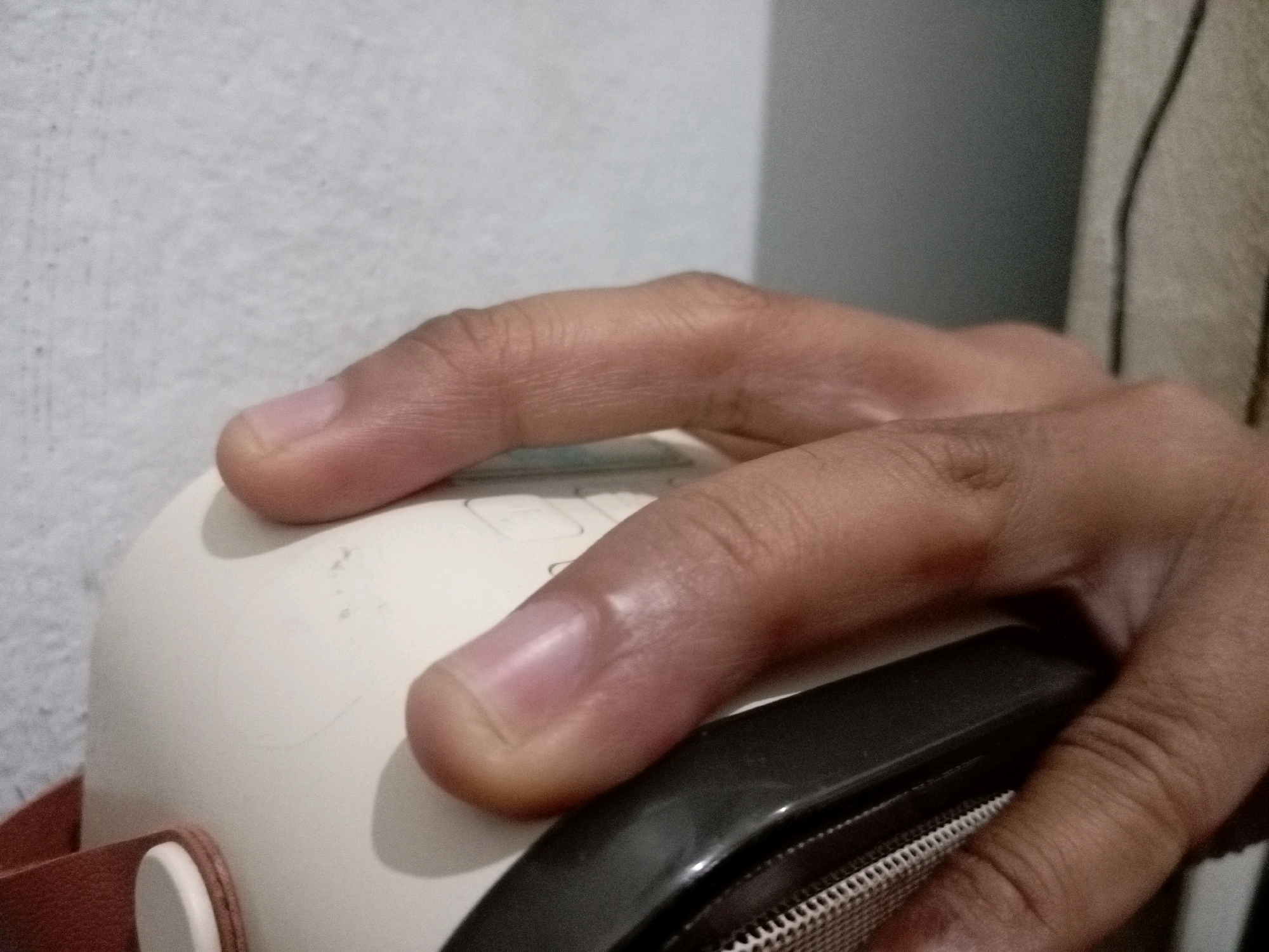 A close-up, slightly overexposed, indoor shot shows a person's hand, with fingers extended and resting on a white device. The hand is positioned in the center of the frame, partially obscuring the white object and a darker, textured element in the foreground. The background consists of a light-colored textured wall on the left and a darker, vertically striped fabric on the right. No specific location in Batam, Indonesia, can be determined from the visual information.