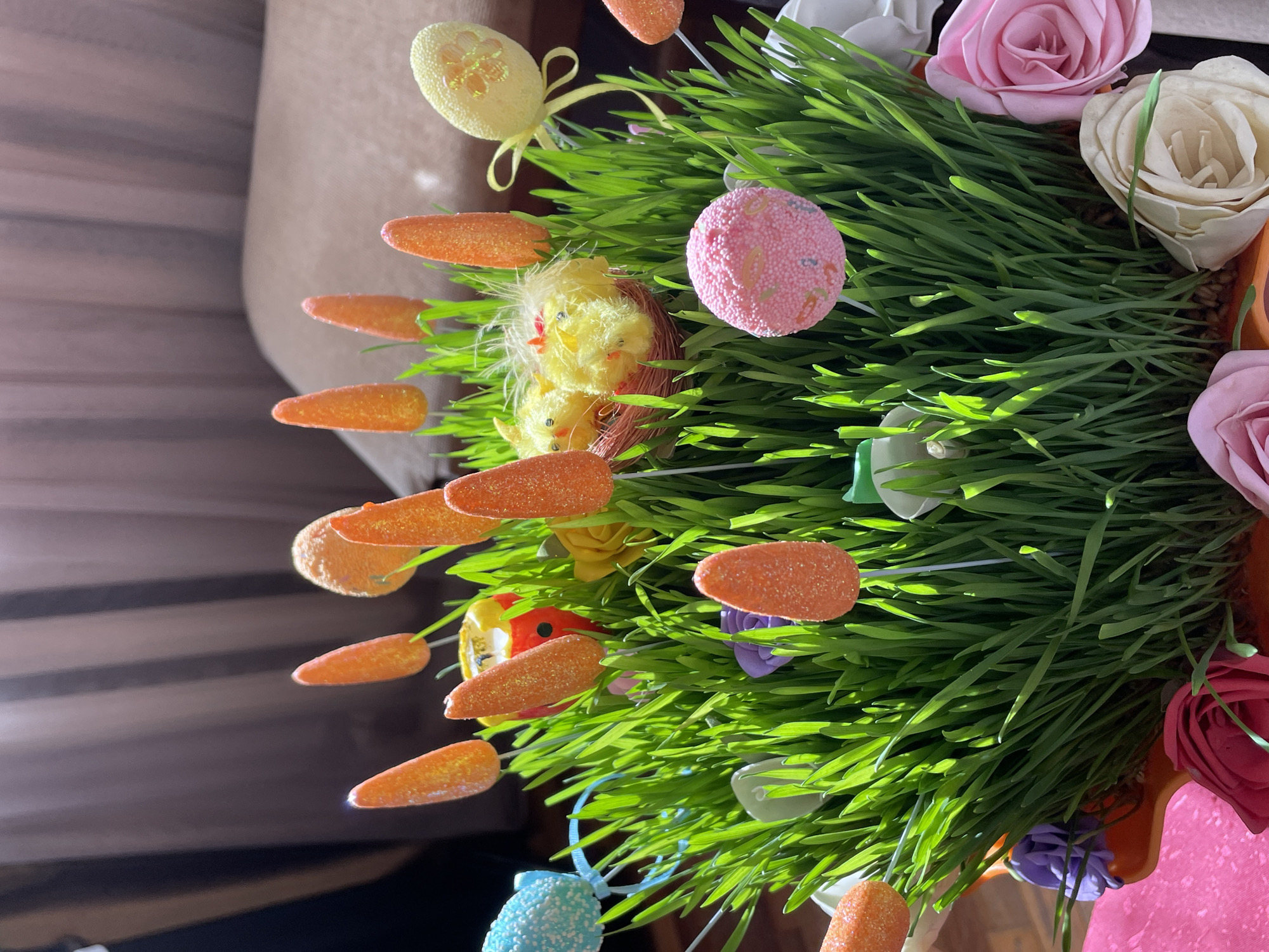 A close-up shot displays an Easter-themed arrangement of green grass, adorned with artificial carrots, roses, and decorative eggs. A pair of small, yellow chicks nestle among the grass blades, surrounded by other colorful Easter decorations. The scene is brightly lit by sunlight. The image provides no discernible location in Aratashen, Armenia.