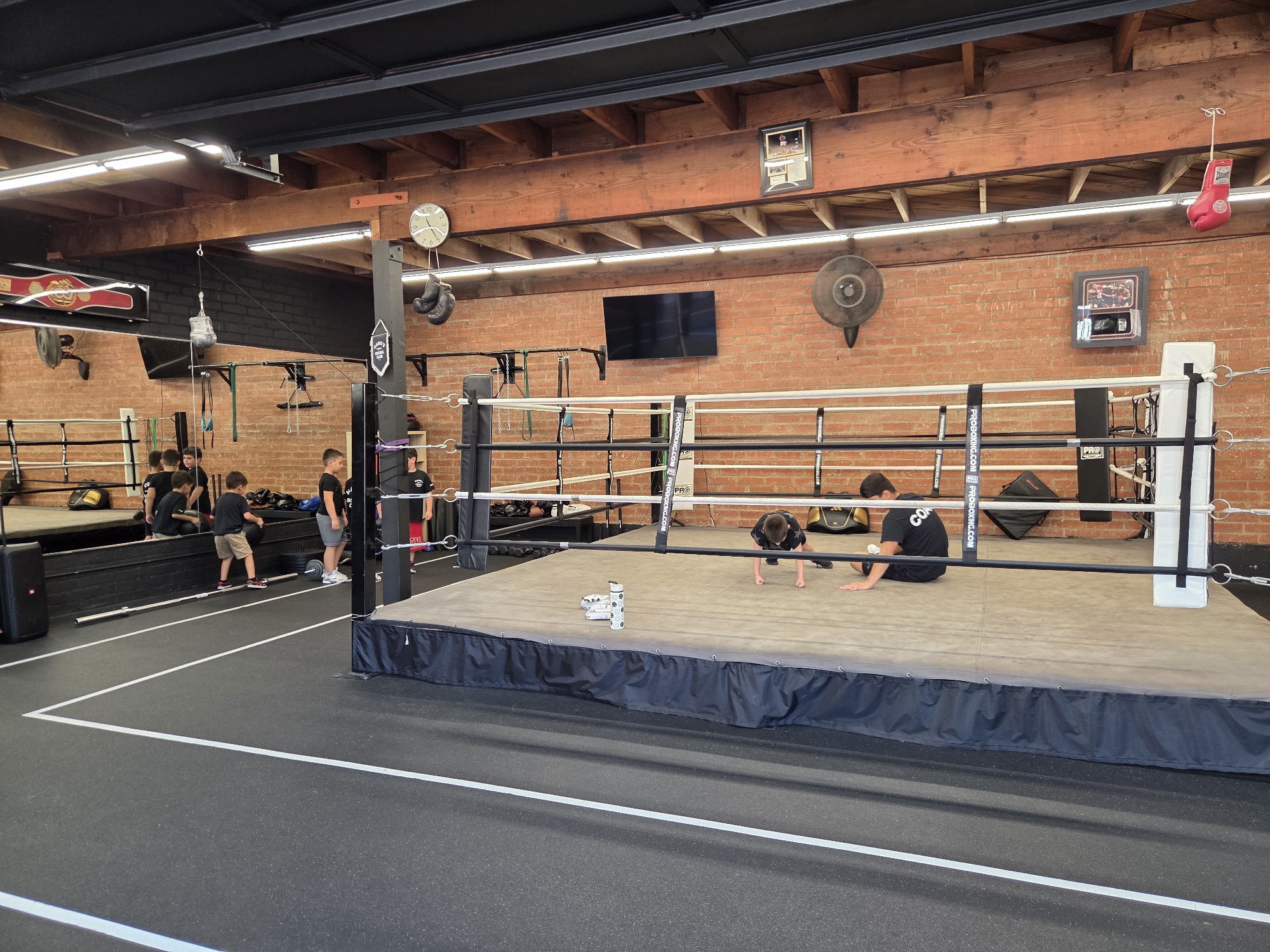 The image depicts an indoor boxing gym in Glendale, United States of America. In the foreground, a boxing ring with a beige mat is centrally located. Two young boys are visible on the boxing mat; one is in a push-up position and the other is seated. To the left of the ring, a group of young boys are gathered. In the background, there are brick walls, exposed wooden beams on the ceiling, and various boxing-related equipment such as punching bags, gloves, and a clock. Lighting fixtures are installed on the ceiling.
