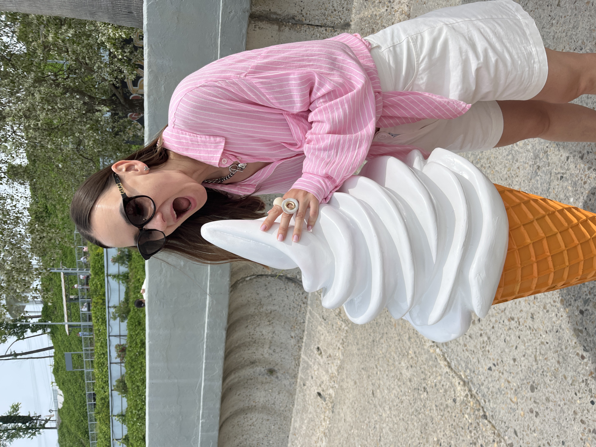 A woman in a pink striped shirt and white shorts is posing with a giant ice cream cone sculpture in Sochi, Russia. The woman is wearing sunglasses and has her mouth open in a surprised expression as she leans towards the ice cream. The ice cream cone is white with an orange waffle cone base. The background shows a paved walkway, a grey concrete wall, and a lush green hillside with trees and a few people in the distance. The sky is overcast.